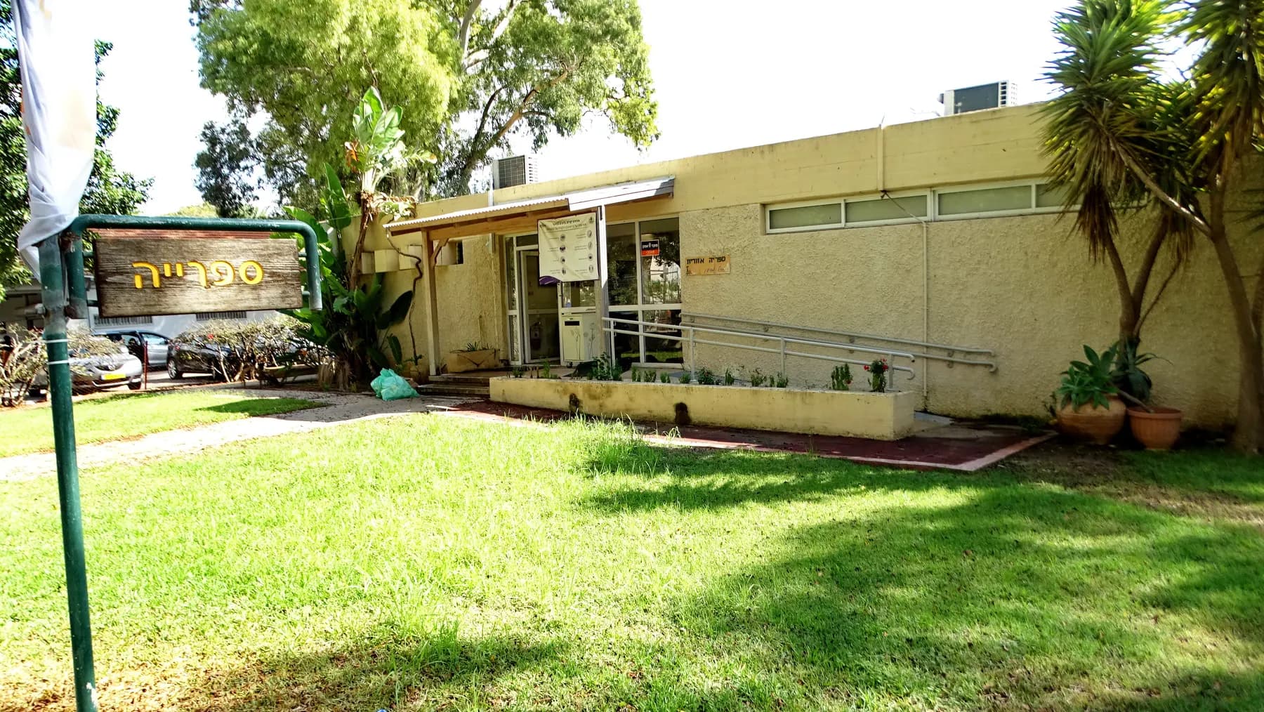 A low yellow library building behind a green lawn in Bnei Atarot, with a palm tree and a wooden sign.