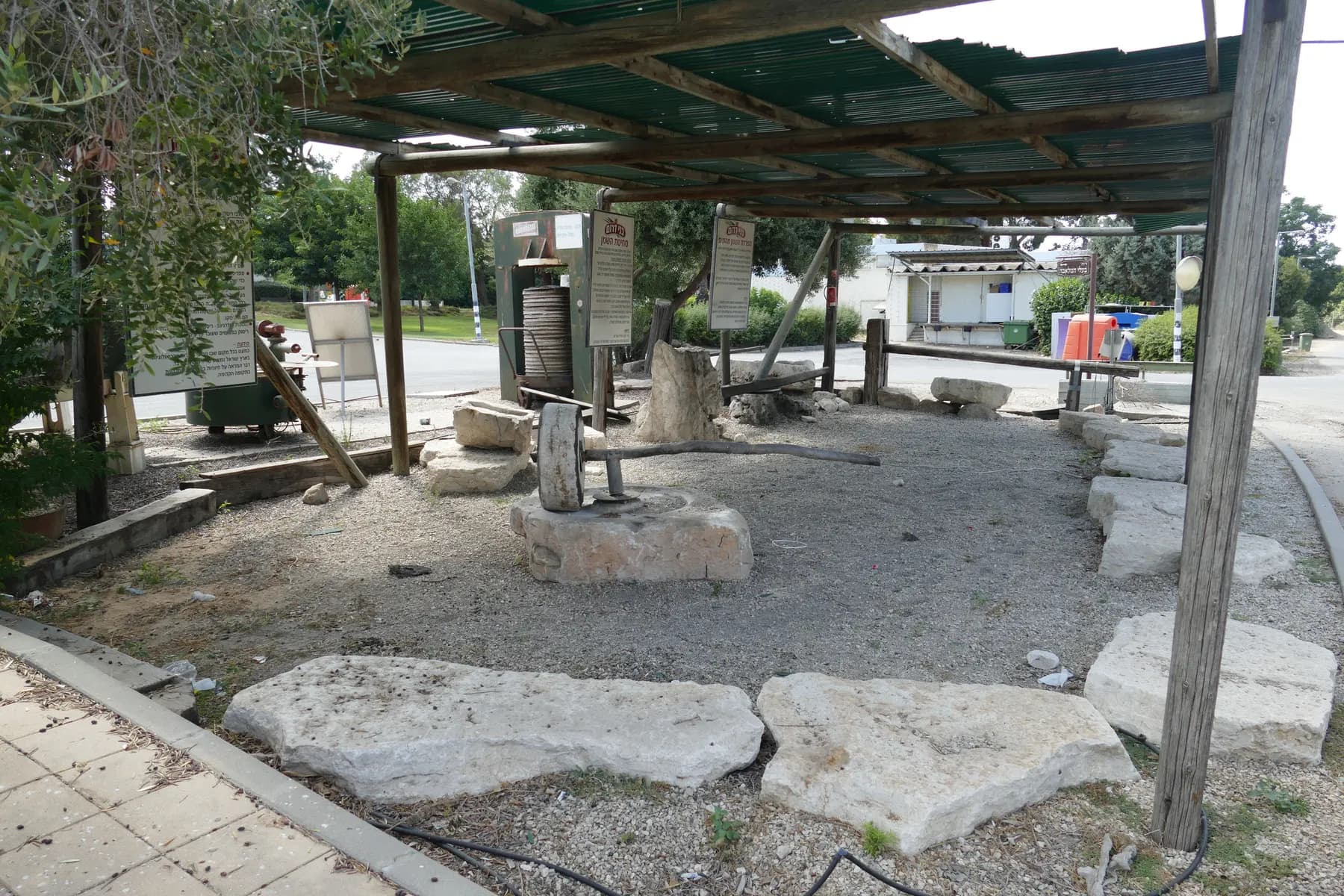 Old olive press stones and a press displayed under a shade roof beside a street in Bnei Darom, with trees and a low building behind.