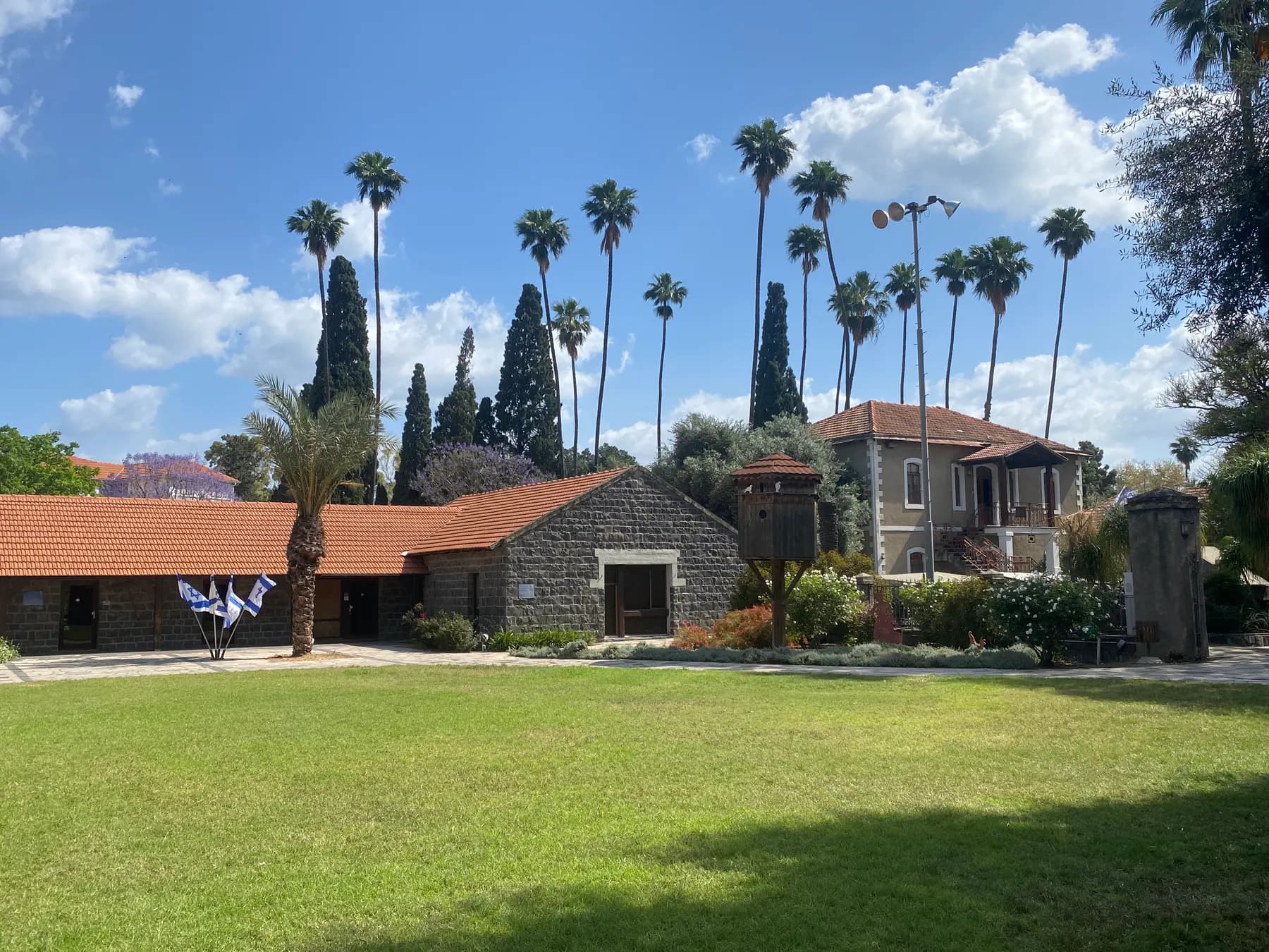 A wide lawn in Degania Alef framed by low basalt-stone buildings with tiled roofs, tall palm trees and a two-storey house.