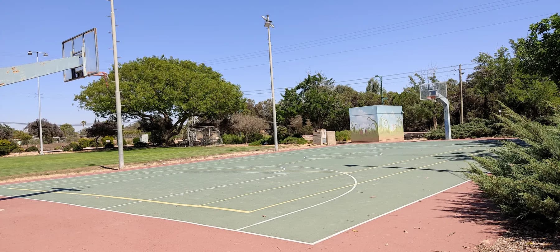 A basketball court with a painted structure at its edge and shade trees behind it in Dekel.