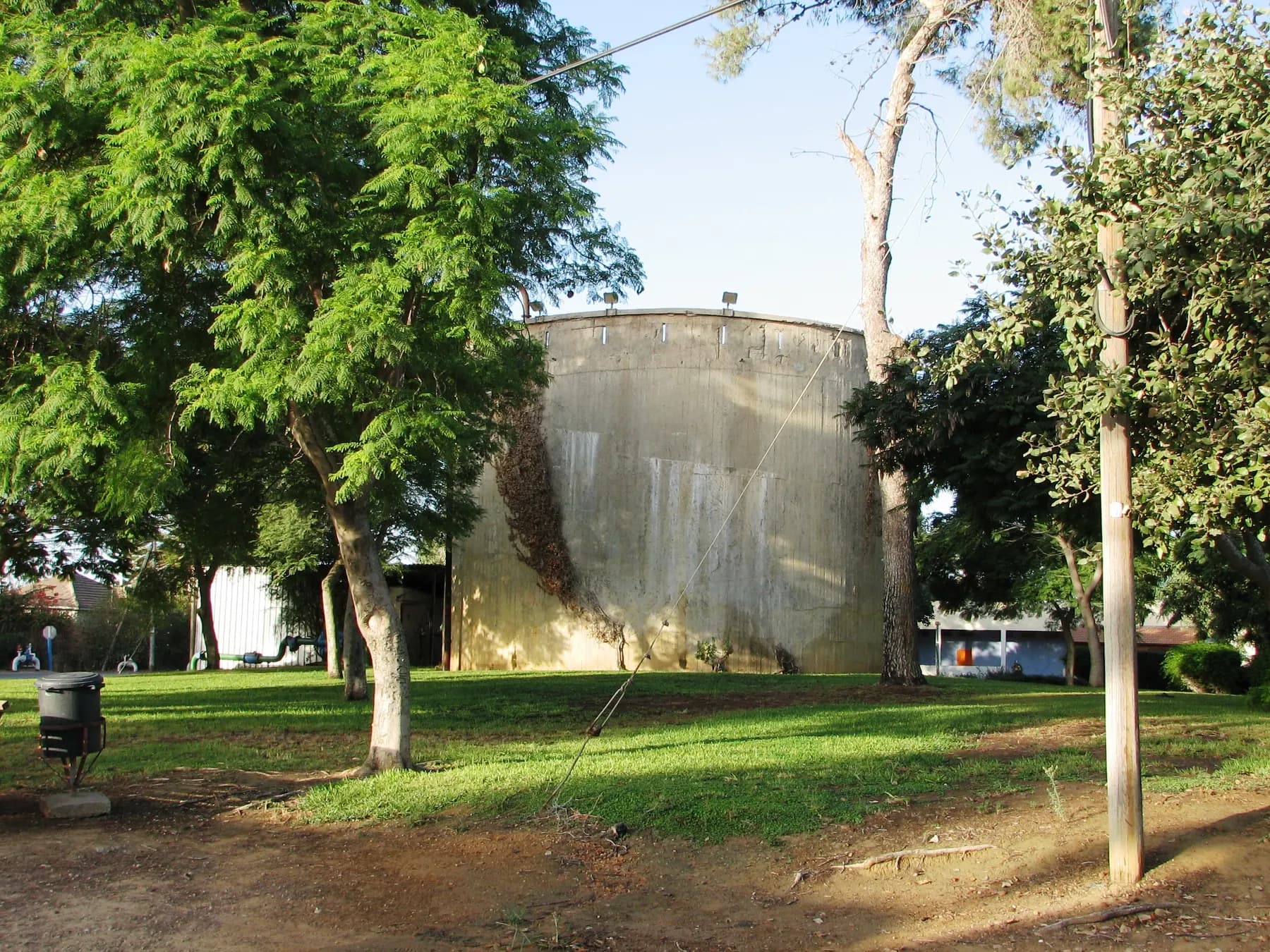 A large round concrete water tank among leafy trees on a lawn in Ein Iron, with low buildings behind it.