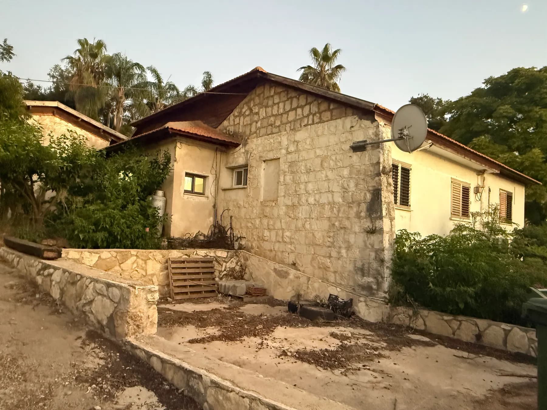An old stone house with a tiled roof and a satellite dish in Ein Ya'akov, above low stone terrace walls, with palms and trees behind.
