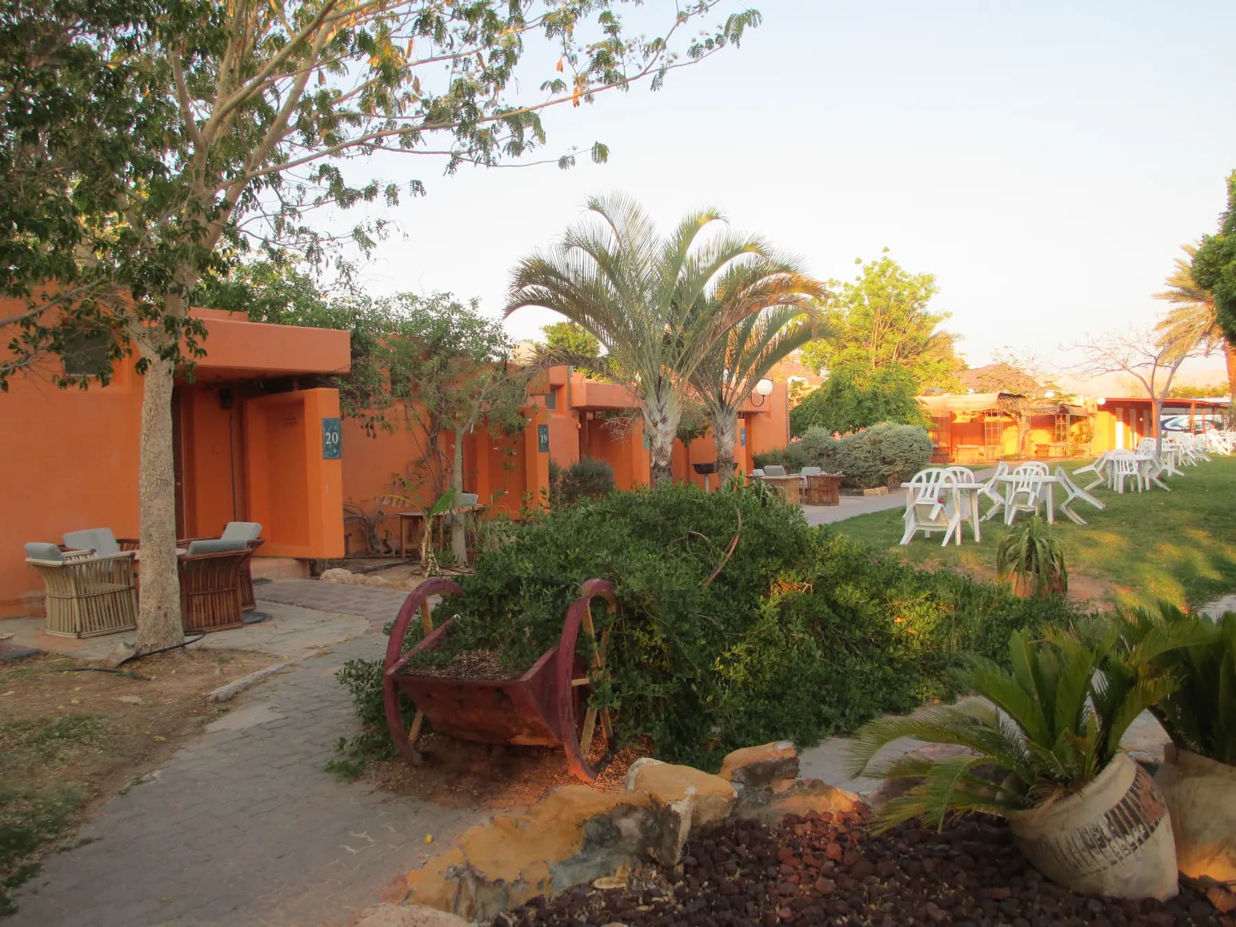 Orange single-storey buildings along a paved path in Elifaz, with palm trees, an old wooden cart full of shrubs and white chairs on a lawn.