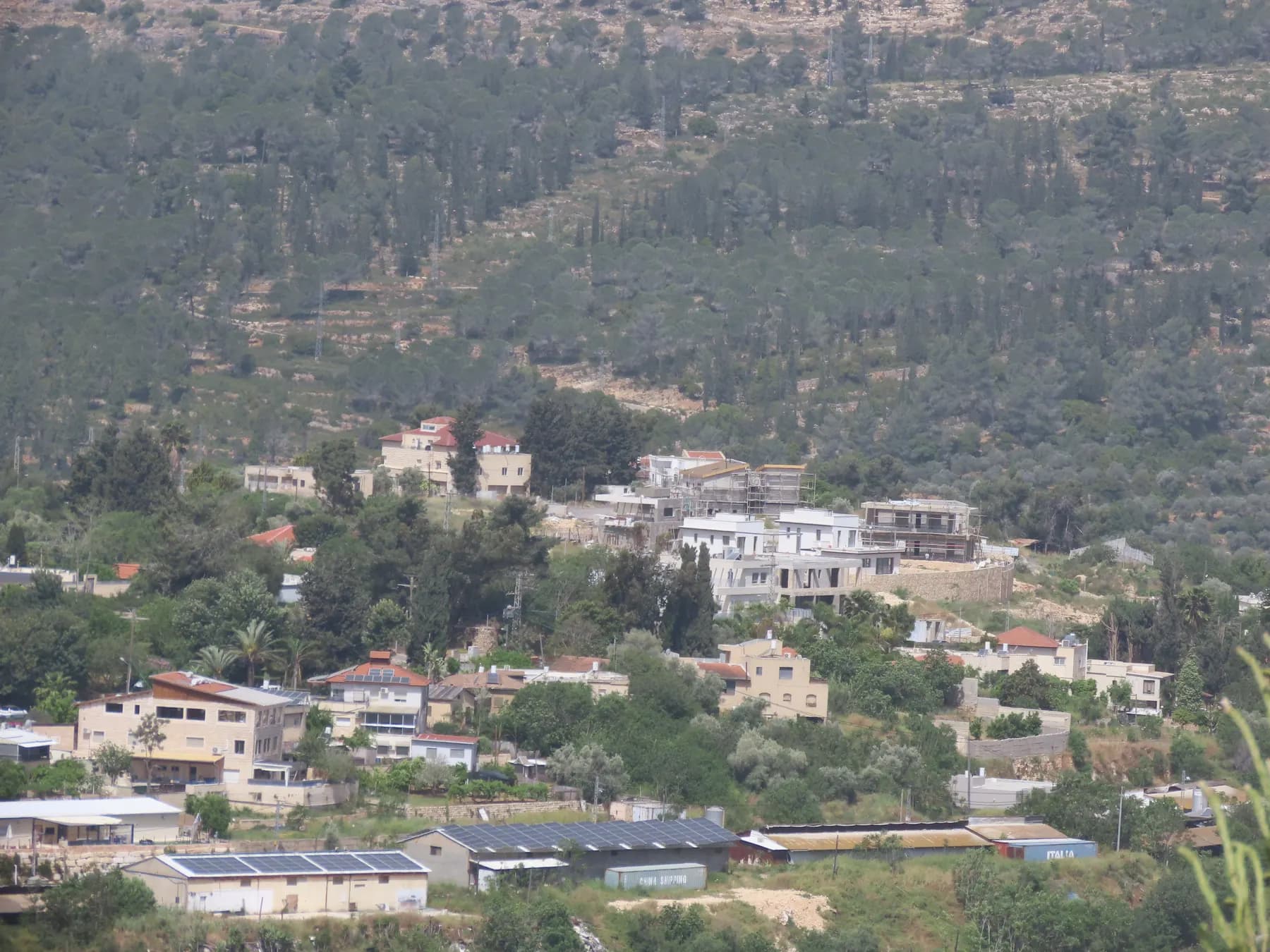 Houses of Even Sapir stepping up a hillside among trees, with farm sheds topped with solar panels below and a pine-covered slope behind.