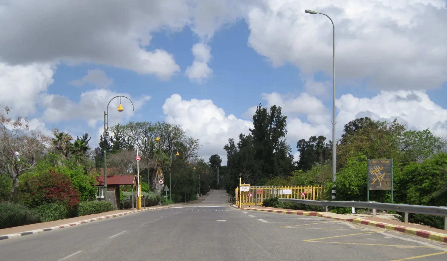The entrance road into Ginaton, with a yellow gate, street lamps and tall trees under a cloudy sky.