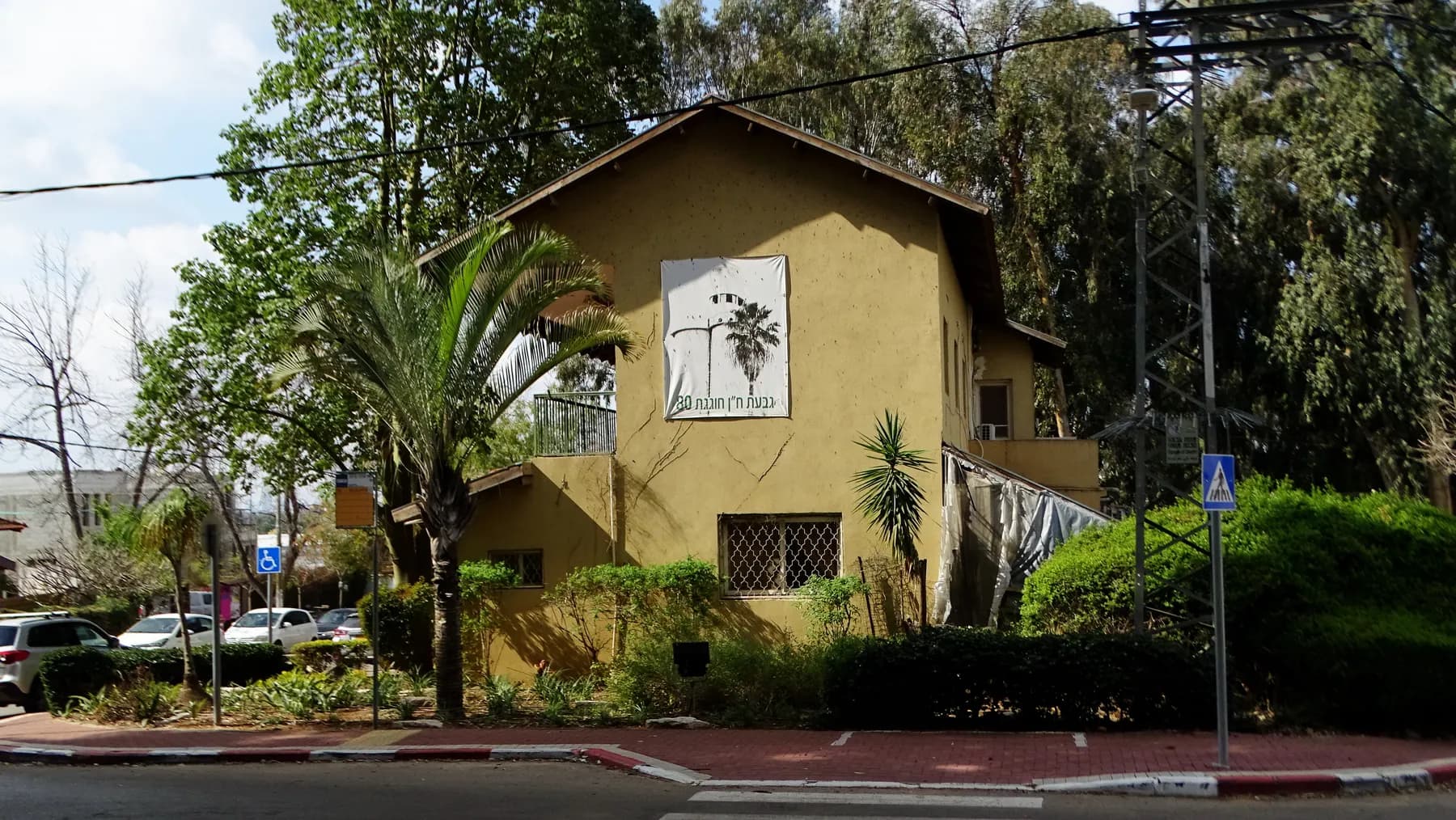 The two-storey secretariat building of Givat Hen, painted yellow with a tiled roof, behind a palm tree and a street with a pedestrian crossing sign.