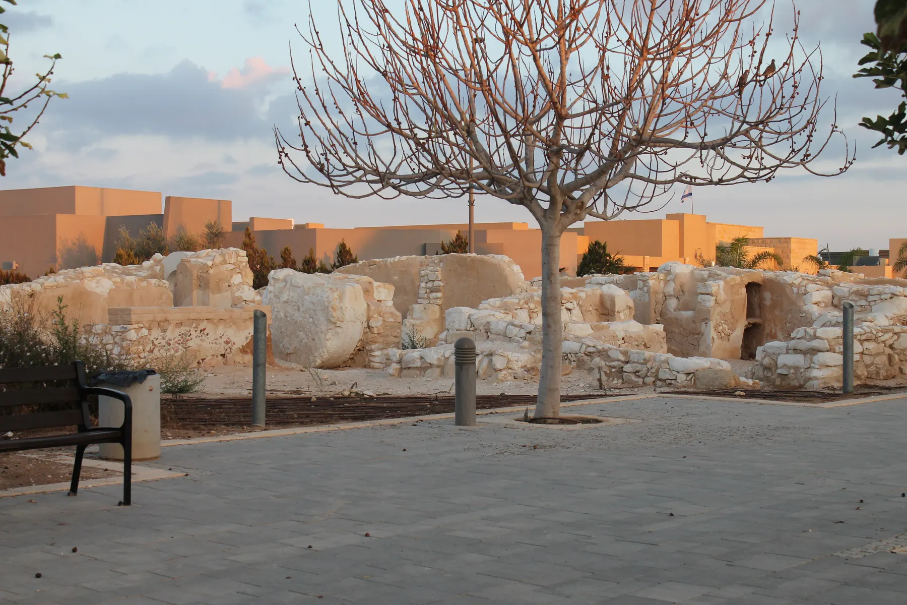Low ancient stone walls in a paved square in Giv'ot Bar, with a bare tree, a bench and houses behind.
