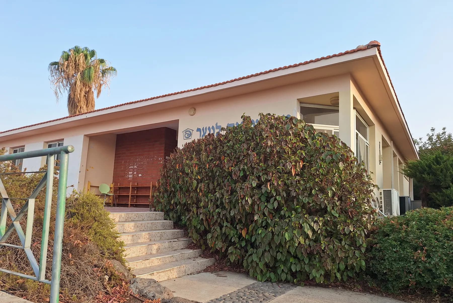 A single-storey building with a tiled roof in Had Nes, reached by a flight of steps beside a large bush, with a palm tree behind.
