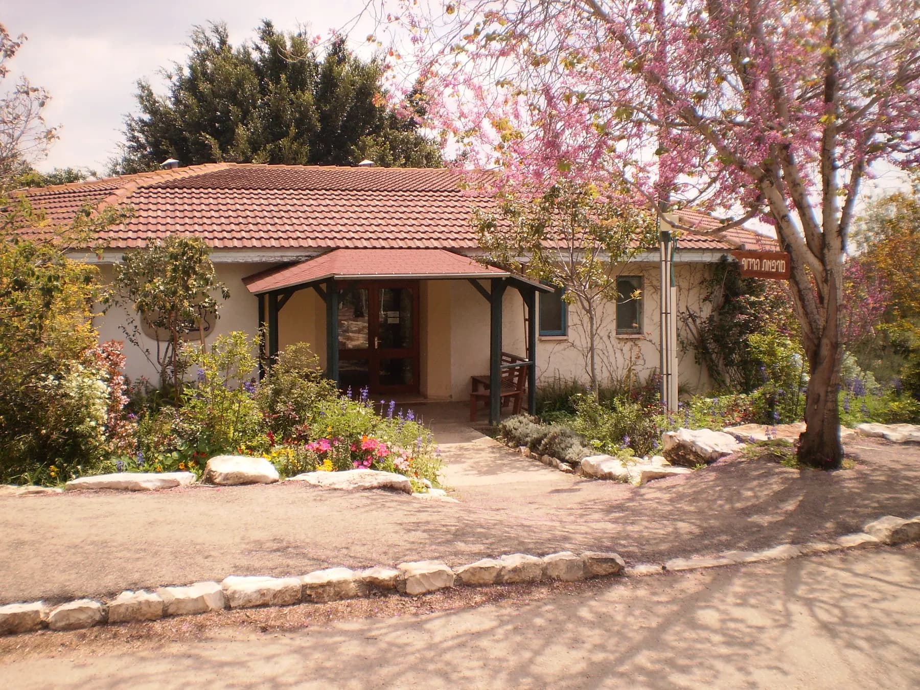 A single-storey building with a red tiled roof and a small porch in Harduf, behind a flower garden edged with stones and a tree in pink blossom.