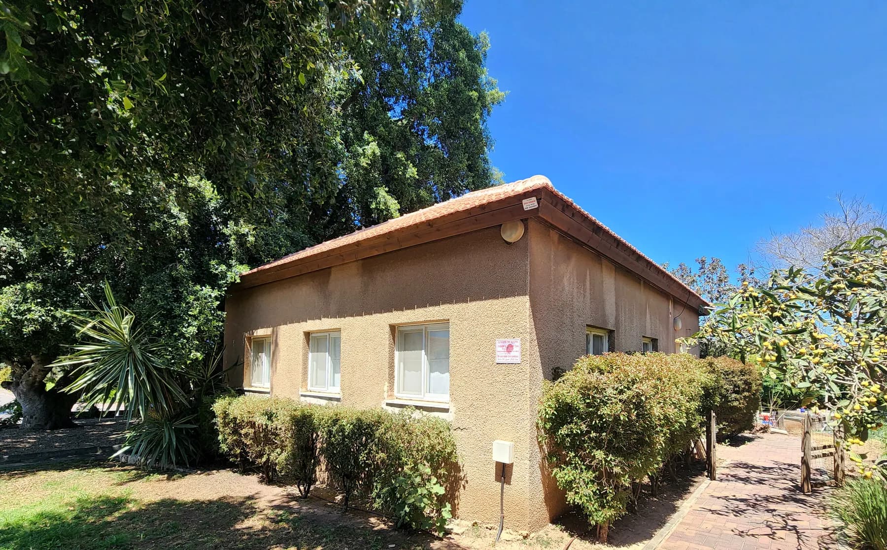 A single-storey kindergarten building with a tiled roof in Hogla, with trimmed hedges along its wall and tall trees beside it under a deep blue sky.