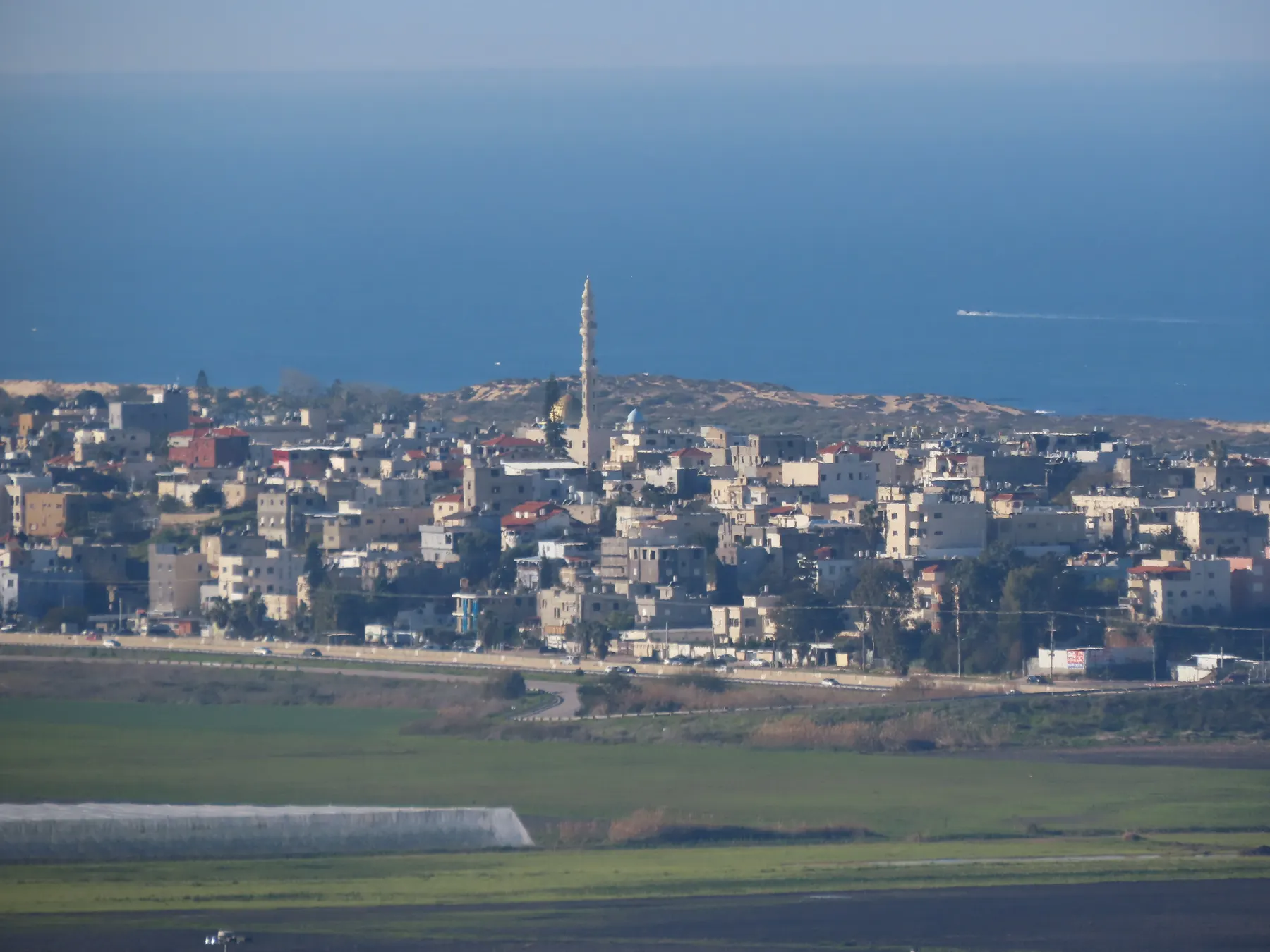 The houses of Jisr az-Zarqa and a tall minaret seen across green fields, with the sea behind.