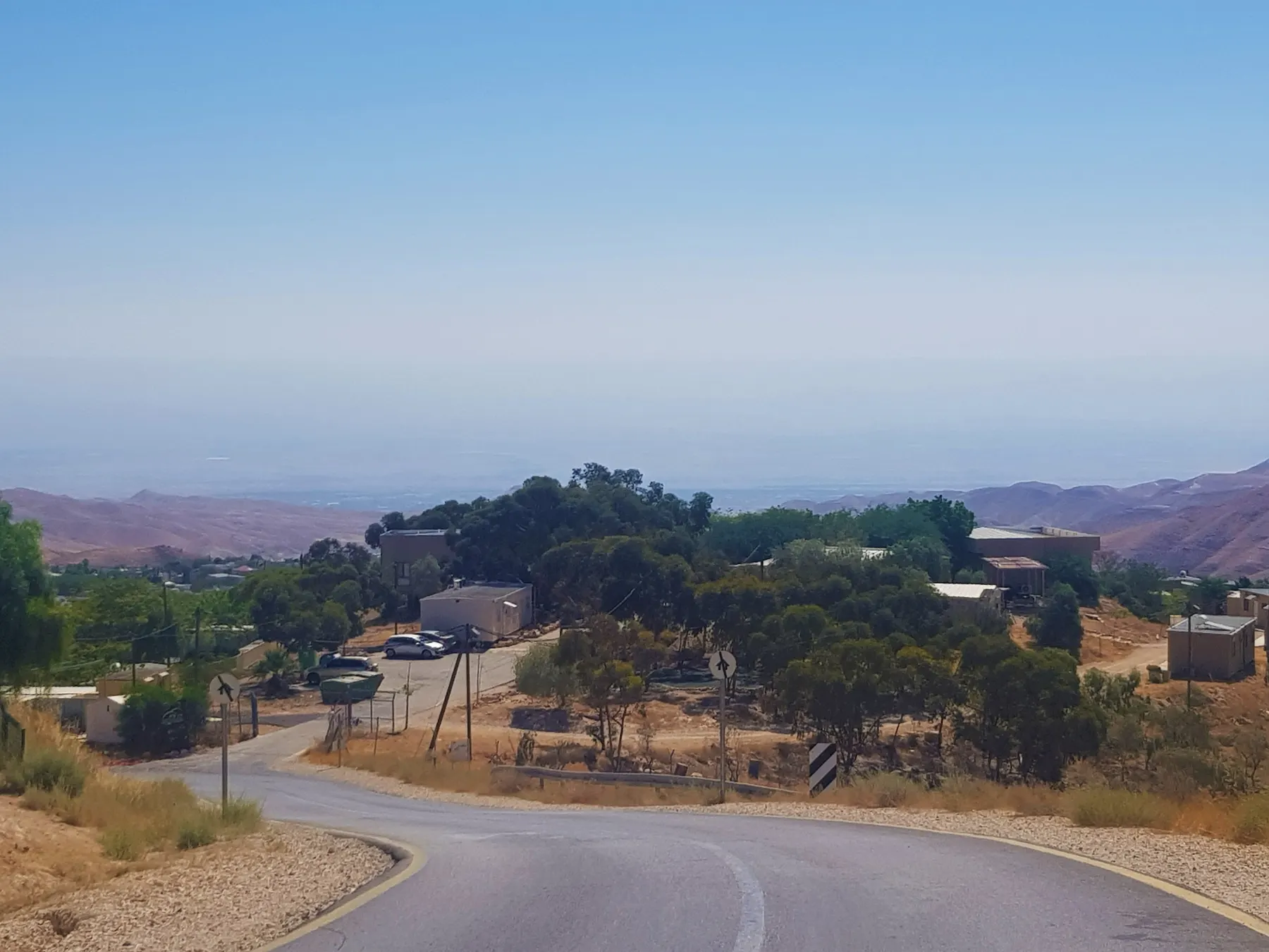 A road winding down into Kfar Adumim, with low buildings among trees and desert hills beyond.