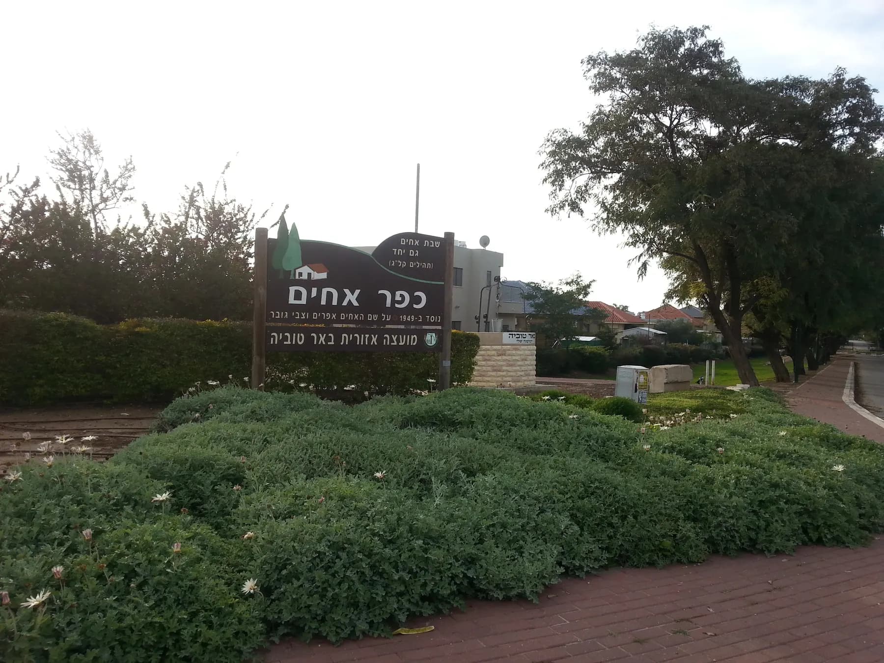 The entrance to Kfar Ahim, with the village sign behind a bed of low shrubs and white flowers, a brick pavement and houses and trees beyond.