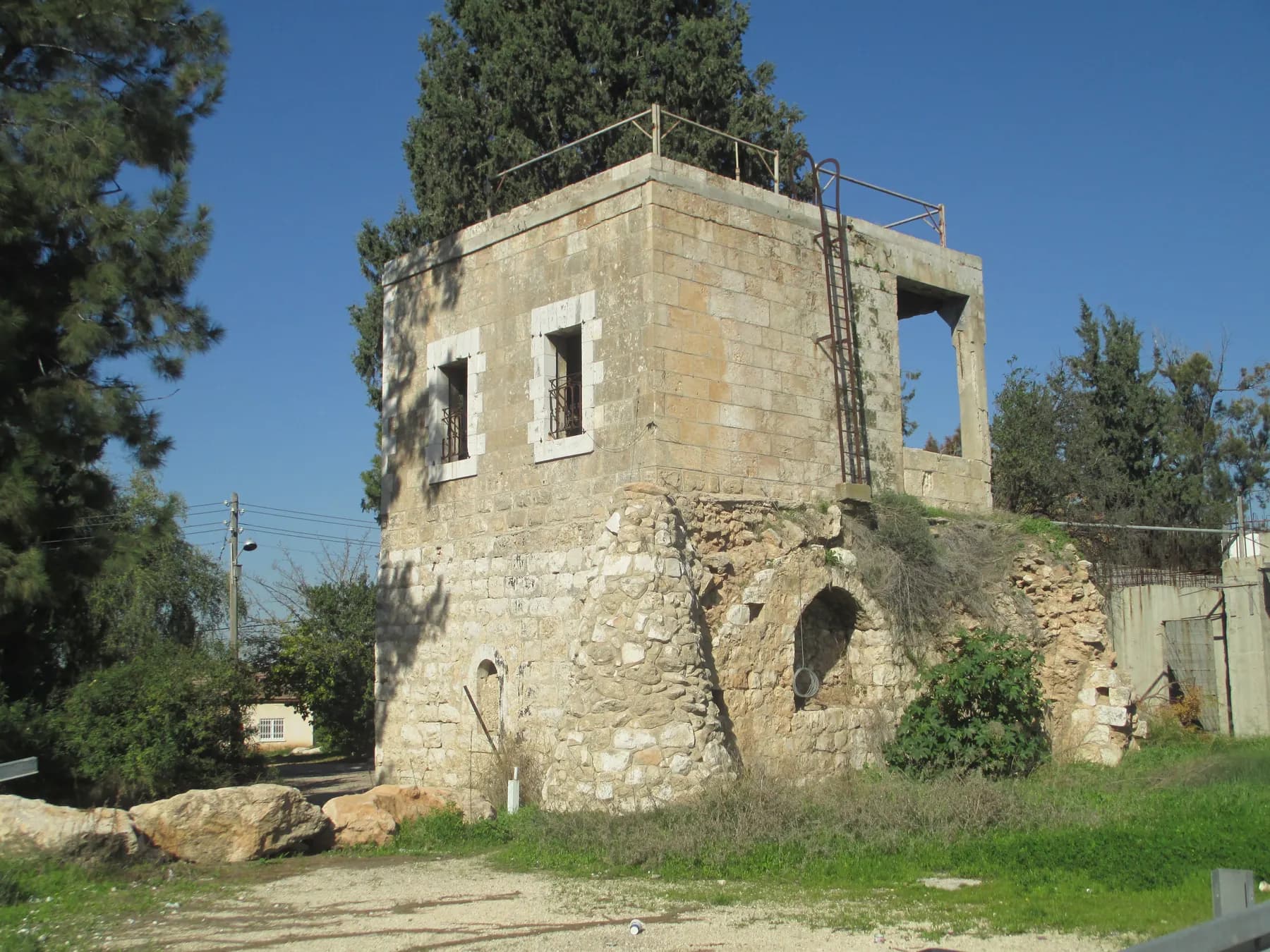 An old two-storey stone building in Kfar Daniel, with an iron ladder up to its flat roof, cypress and pine trees behind it and grass in front.