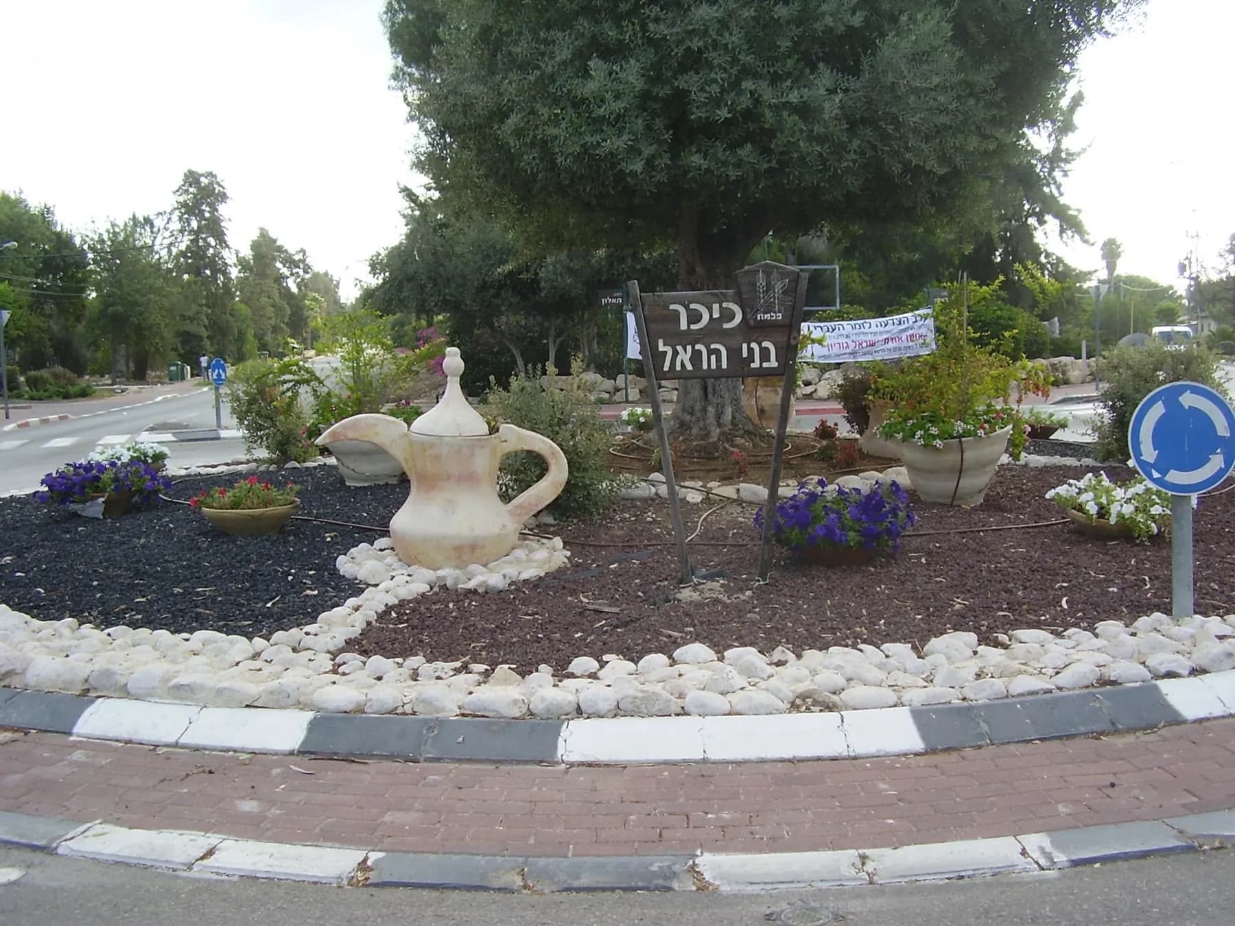A roundabout garden at the entrance to Kfar Truman, with a large coffee pot sculpture, flowers and trees.