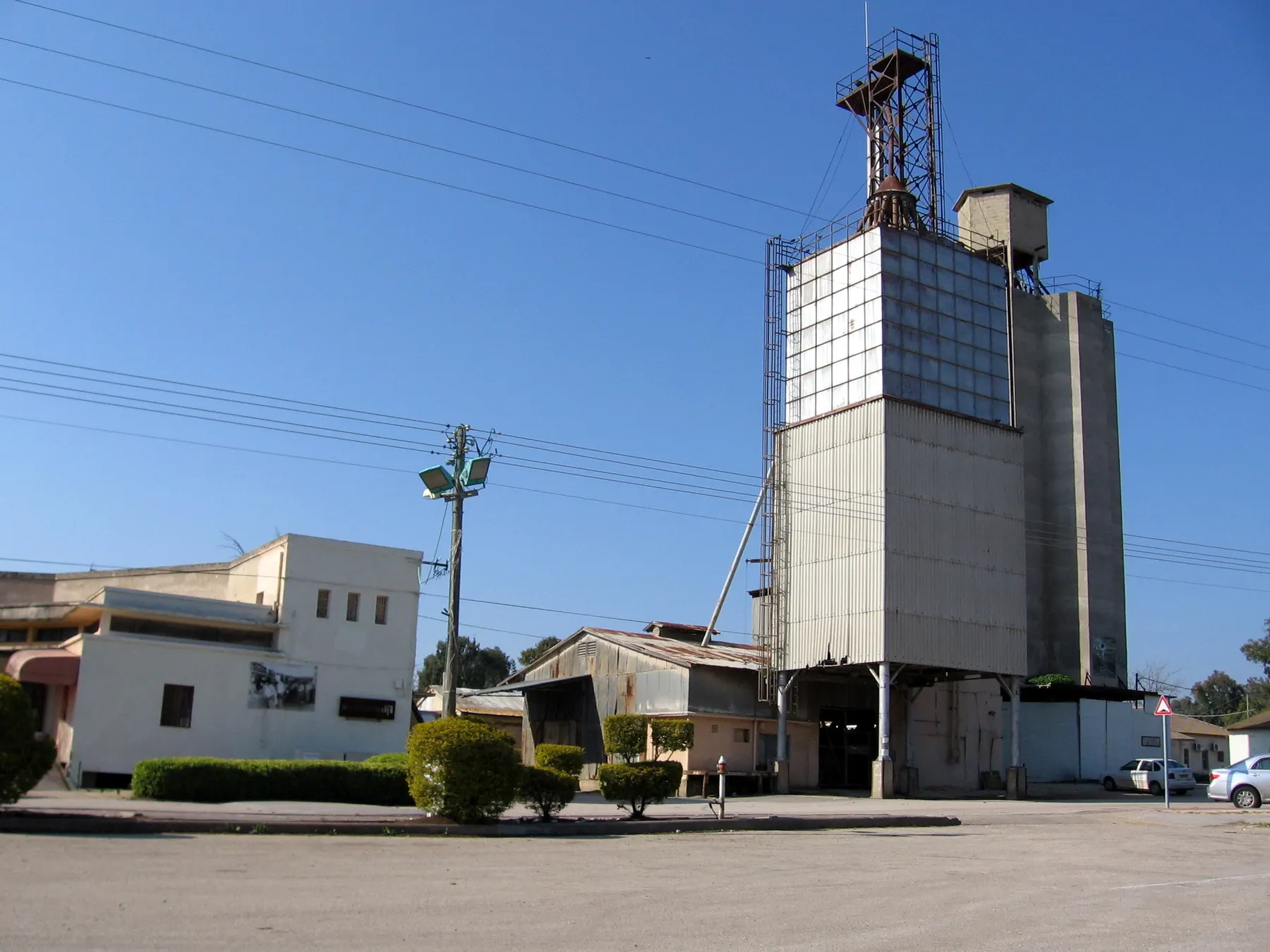 A tall tower clad in metal panels with concrete silos beside it and low farm buildings in front, in Kfar Yehezkel, under a clear blue sky.