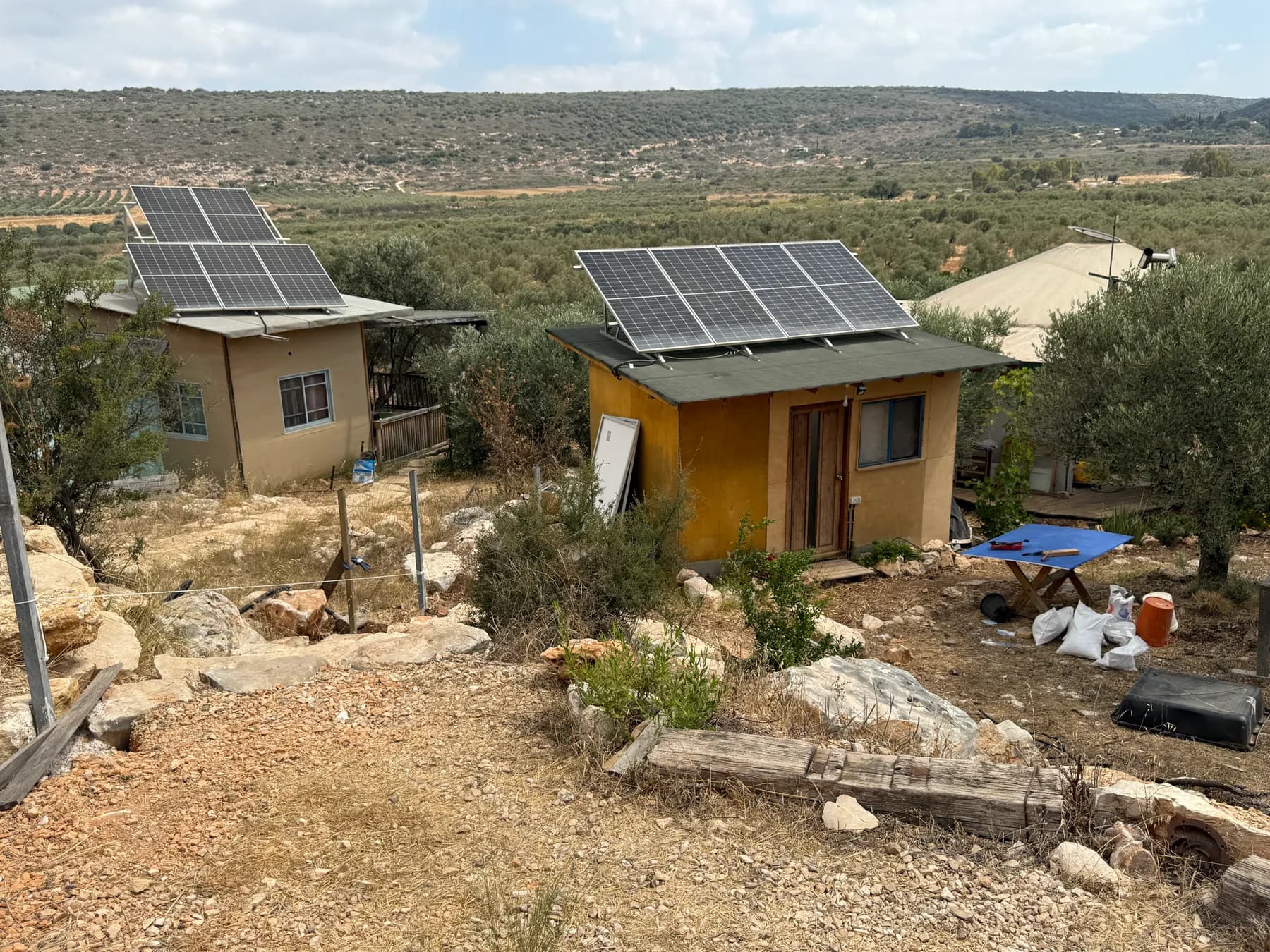 Two small cabins with solar panels on their roofs on a rocky slope in Klil, with olive groves and wooded hills beyond.