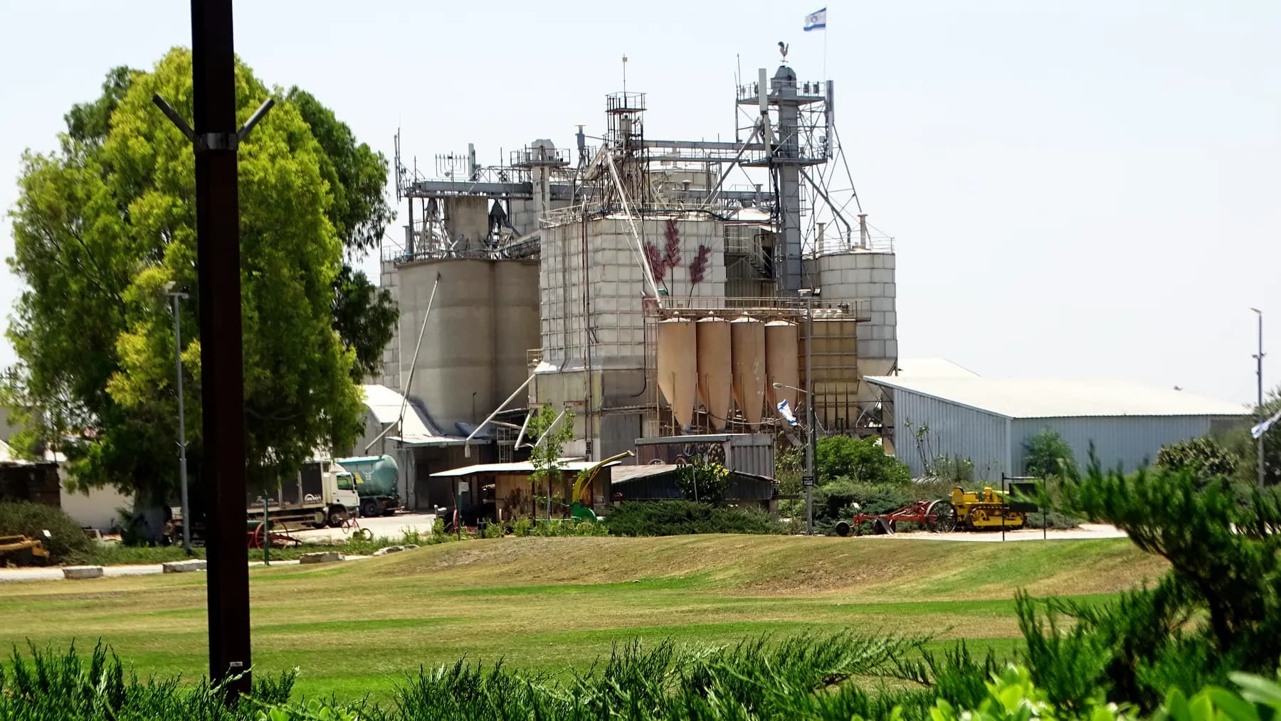 The feed mill of Kvutzat Yavne, a cluster of silos and towers with a flag on top, seen across a lawn with trees on the left.