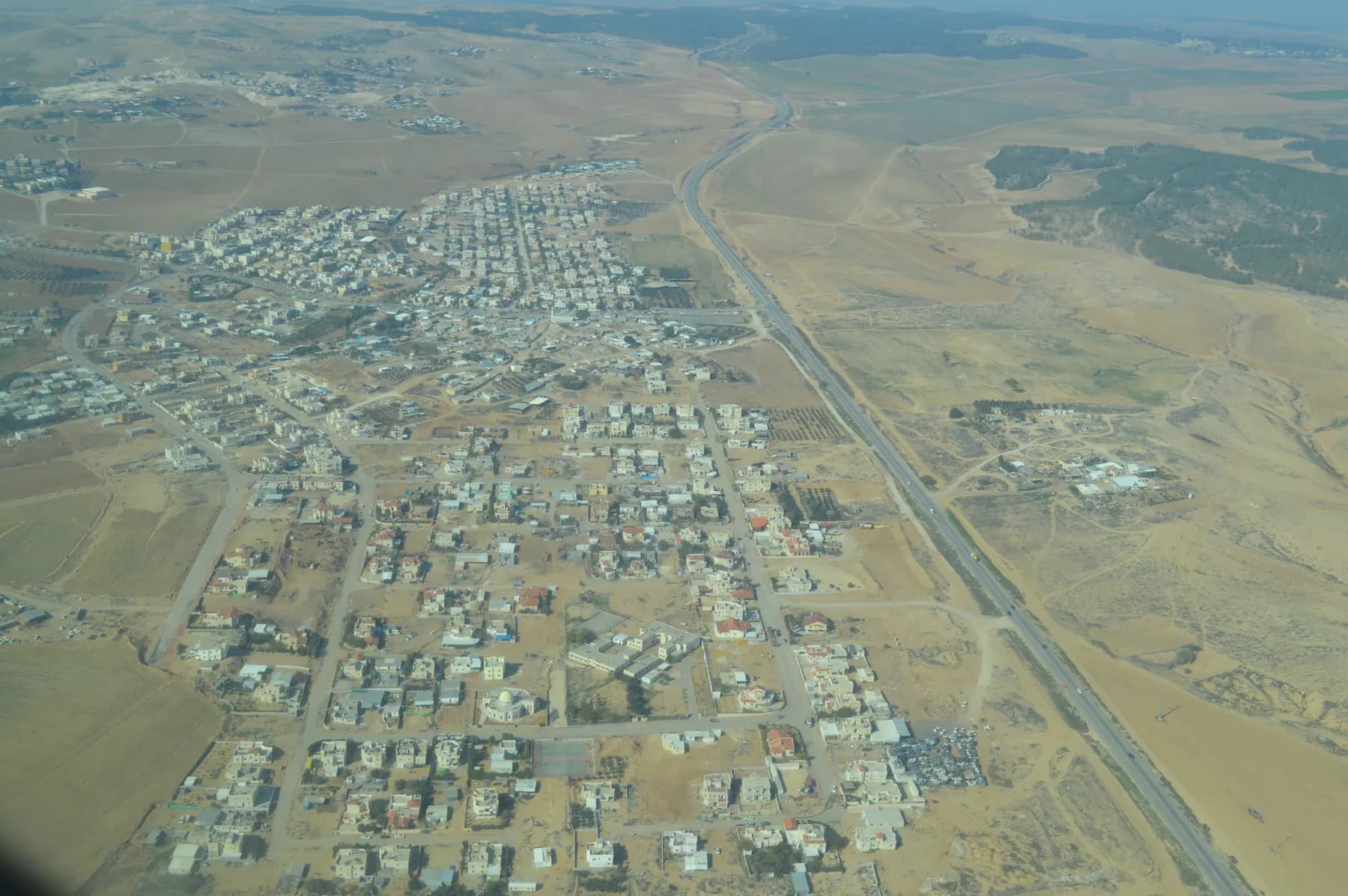 An aerial view of low houses laid out on a grid of streets in Lakiya, beside a highway running through open desert land.