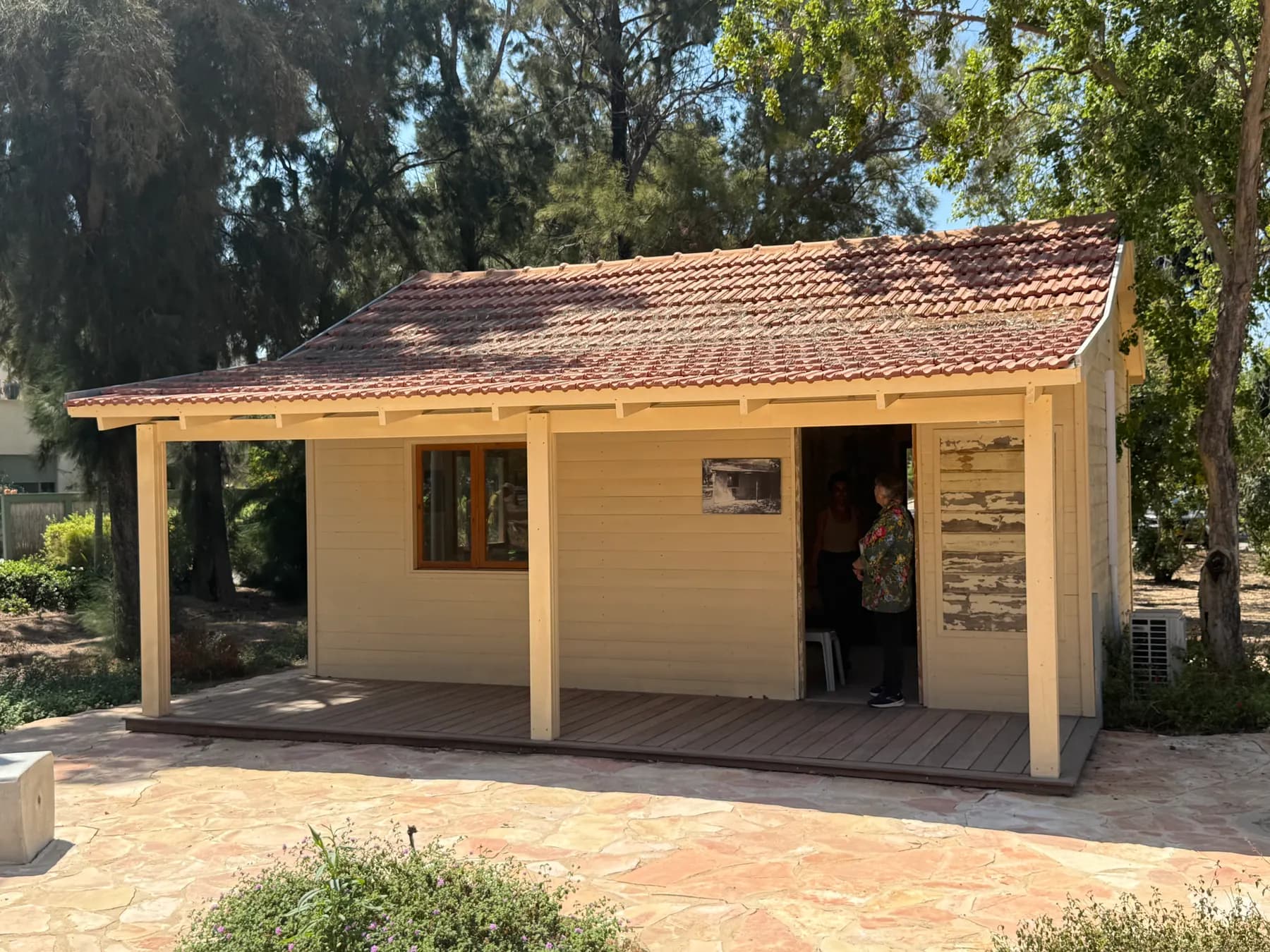 A cream-painted wooden hut with a tiled roof and a covered porch in Lohamei HaGeta'ot, standing on a paved yard among tall trees, with a visitor in the doorway.
