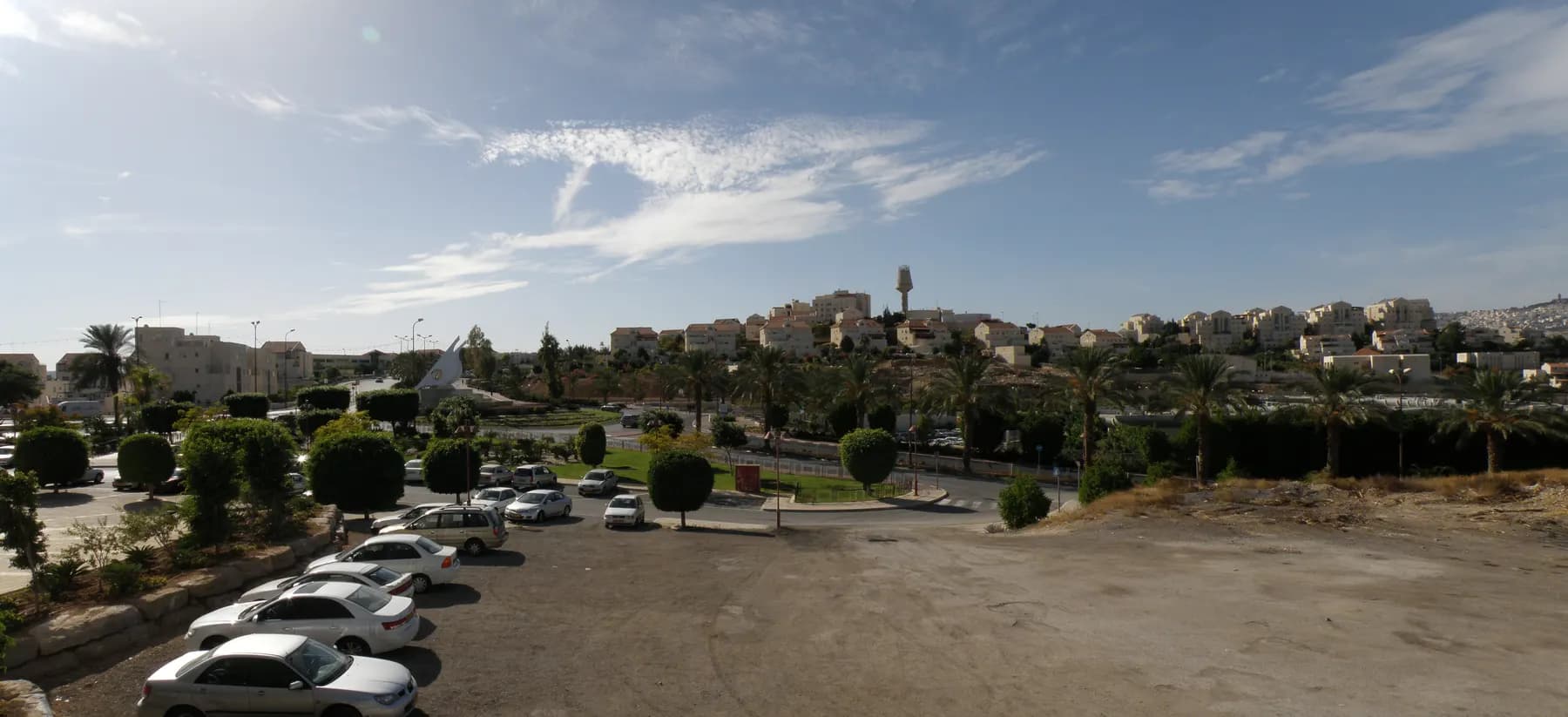 A palm-lined road and car park in Ma'ale Adumim, with houses and a water tower on the hill behind.