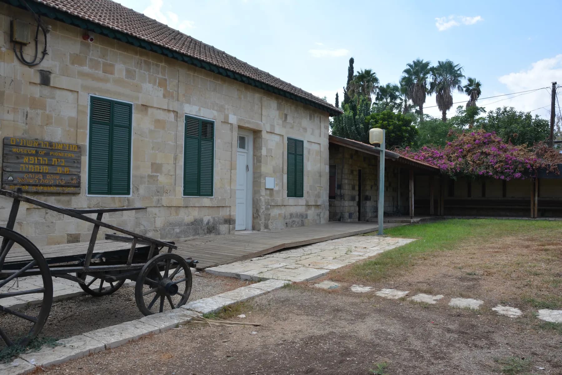 A stone building with green shutters and a tiled roof in Menahemia, with an old wooden cart in front and a long low wing covered in pink bougainvillea beside a lawn.