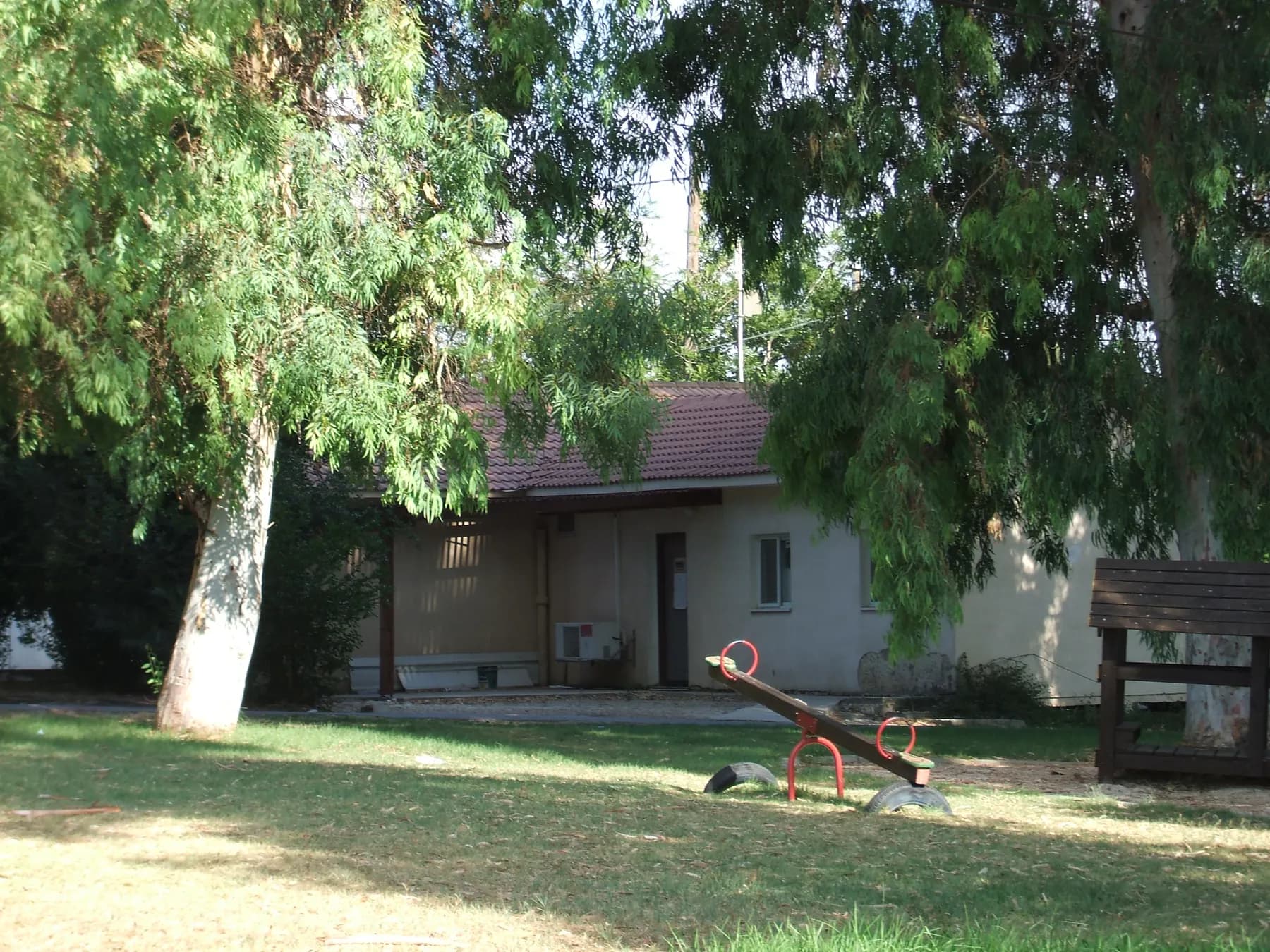 A low house with a tiled roof behind a lawn with a seesaw, shaded by tall trees in Midrakh Oz.