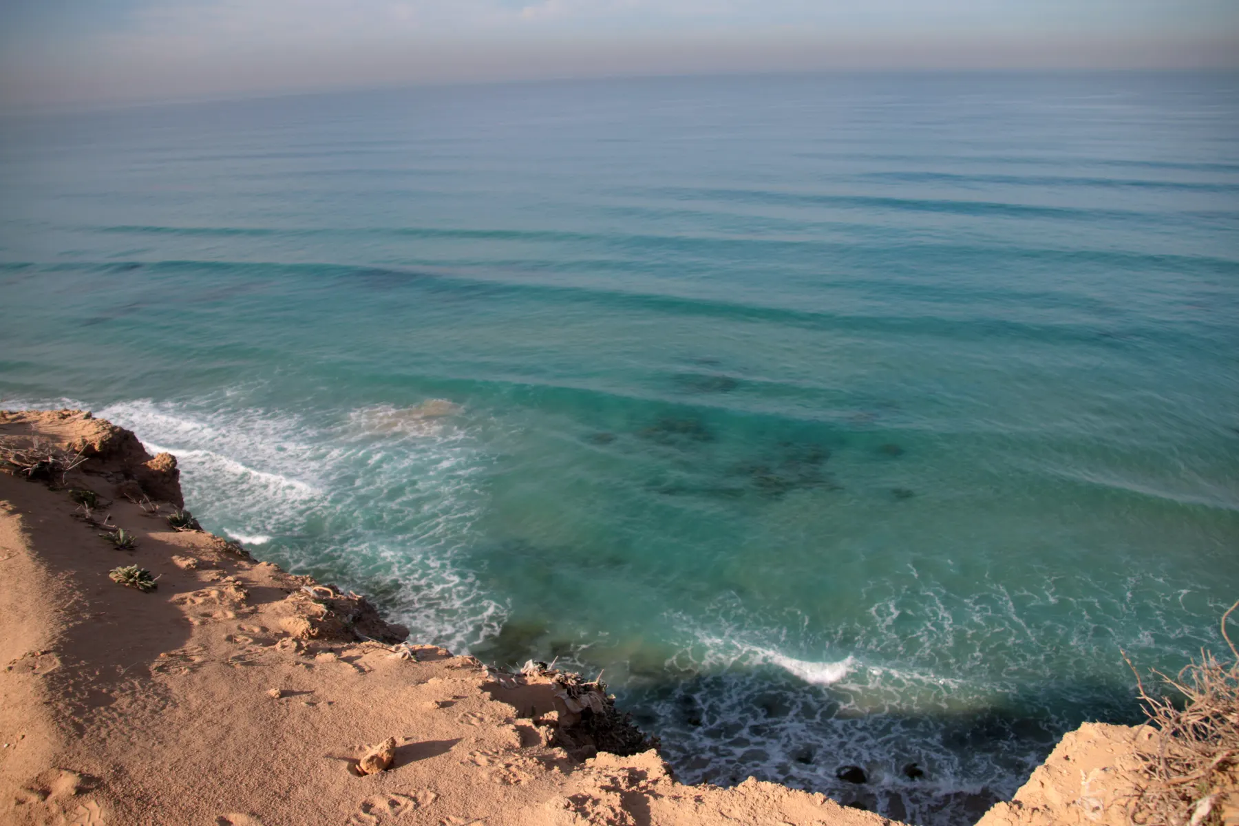A sandstone cliff edge above turquoise sea and small breaking waves on the coast at Ramat Poleg, Netanya