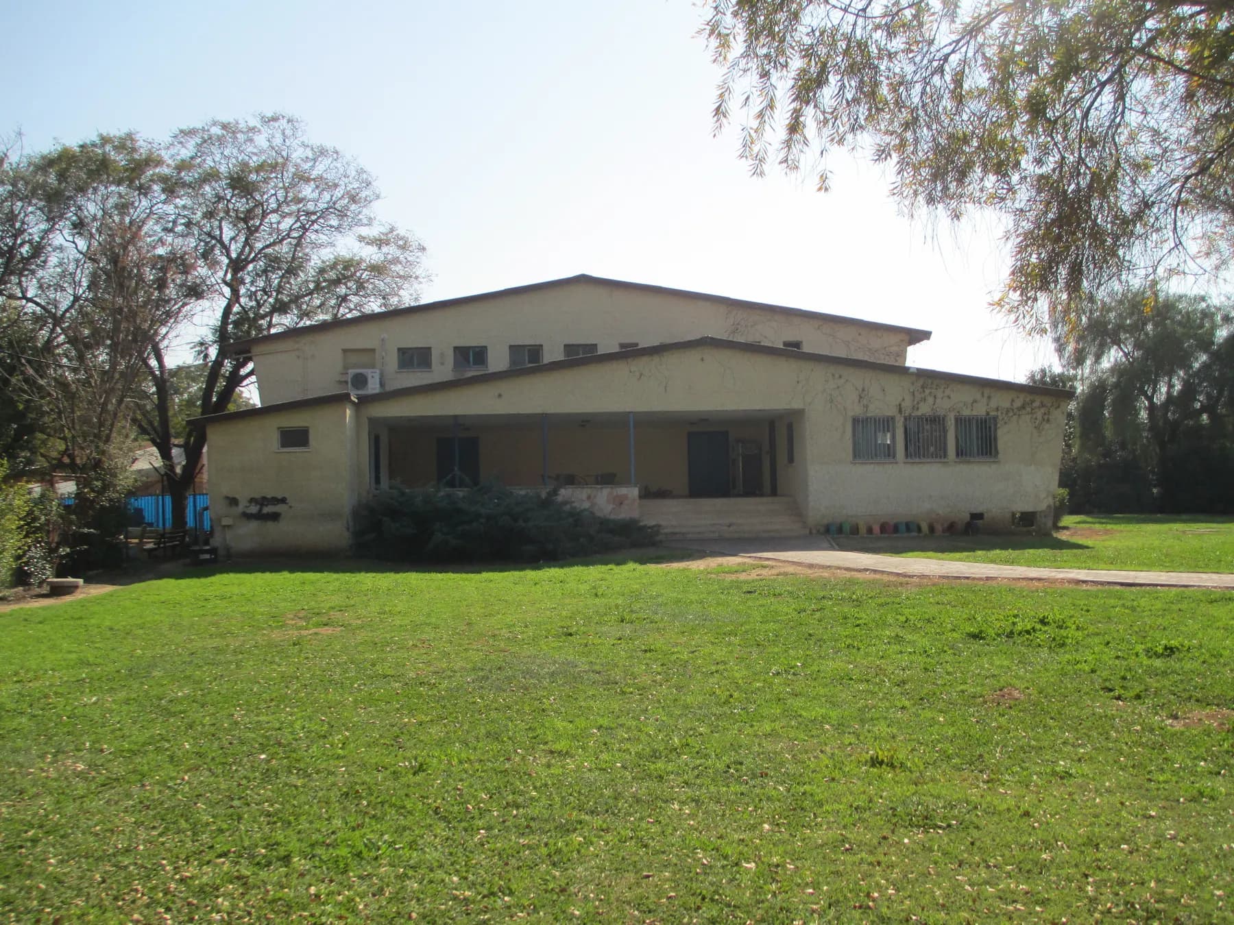 The community hall of Nir Banim, a pale building with a covered entrance and steps, behind a wide lawn with trees on both sides.