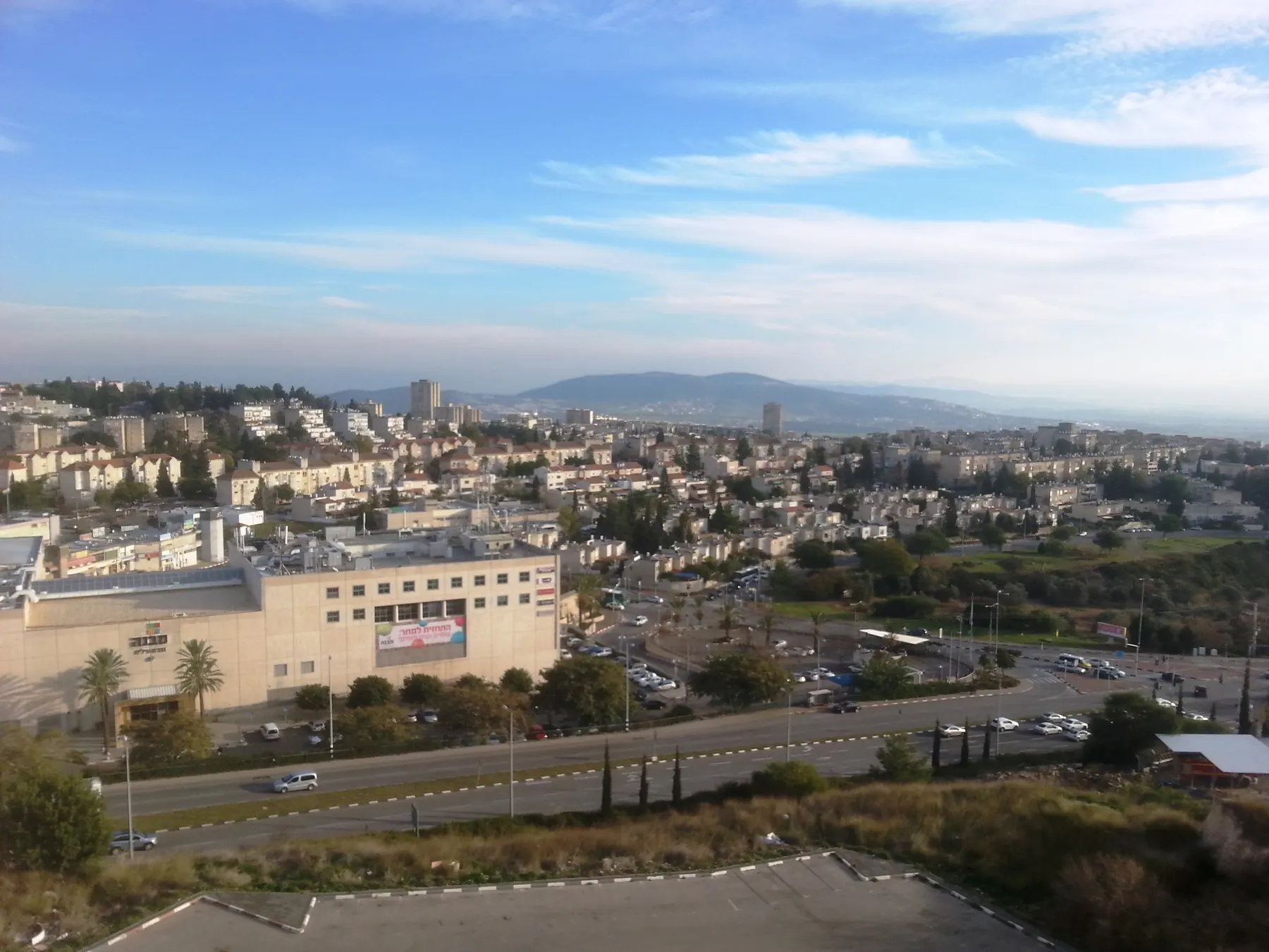 A view over Nof HaGalil from above, with a shopping centre, a main road, houses and hills behind.