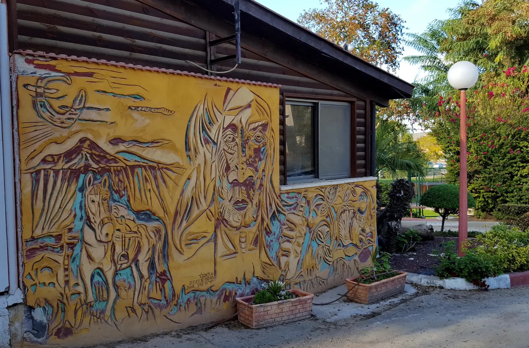 A low building in Or HaNer with a large yellow mural of a face and horses on its wall, beside a globe street lamp, planters and a garden.