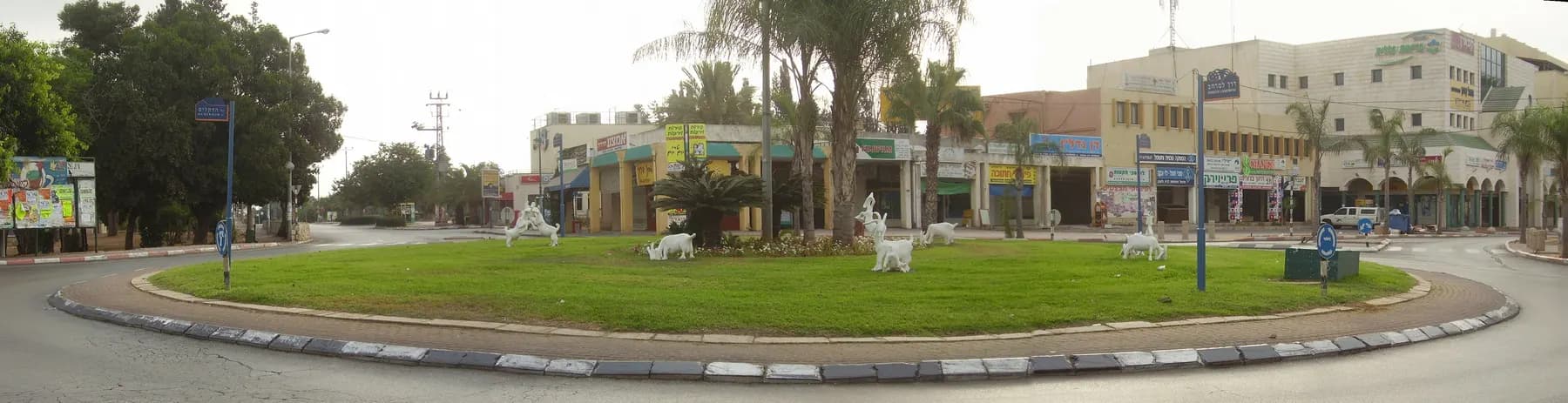 A wide view of HaNasi square in Pardes Hanna: a grass roundabout with white goat sculptures, palm trees and the shops of the town centre behind