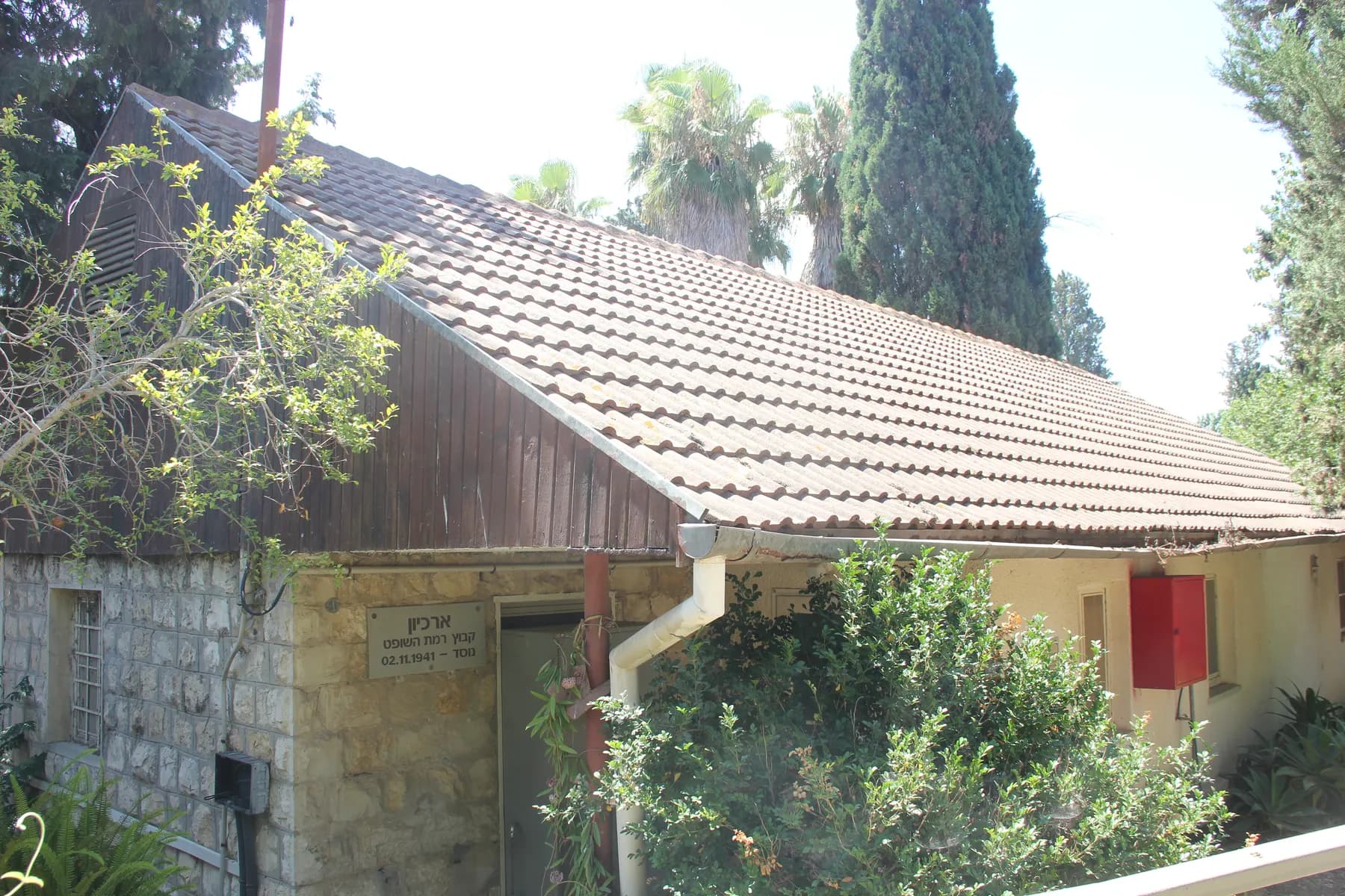 An old building with stone walls and a tiled roof among trees and palms in Ramat HaShofet.