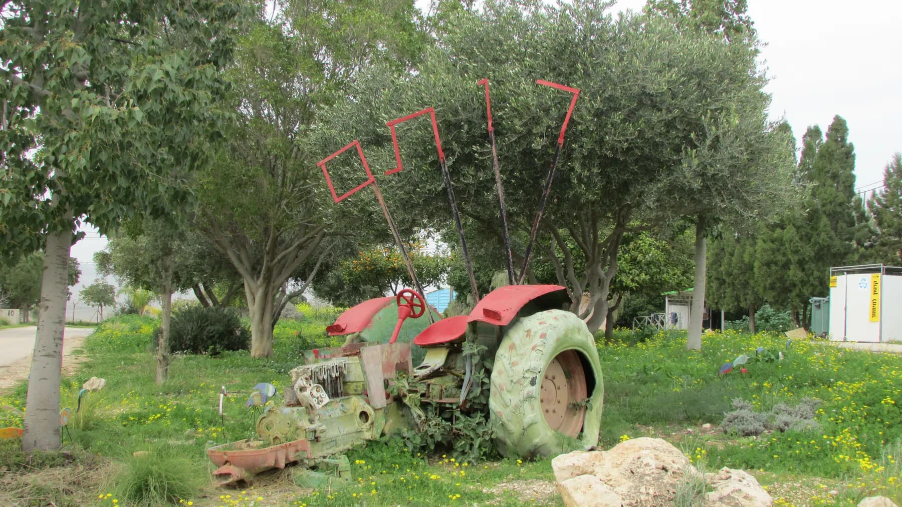 An old tractor painted green and red on a lawn under olive trees in Rotem, with the village's name in red metal letters on poles above it.