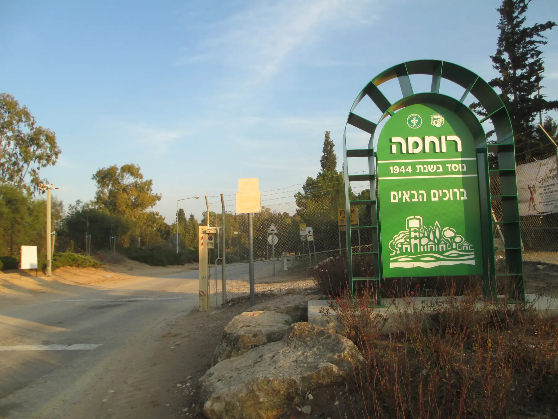 The entrance road into Ruhama, with a green arched welcome sign bearing the kibbutz name, a gate in a wire fence, boulders and eucalyptus trees.
