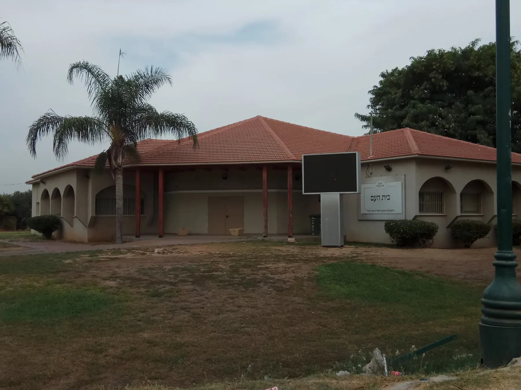 The community hall of Sde Uziyahu, a low building with a red tiled roof, arched windows and a covered porch, behind a lawn with a palm tree.