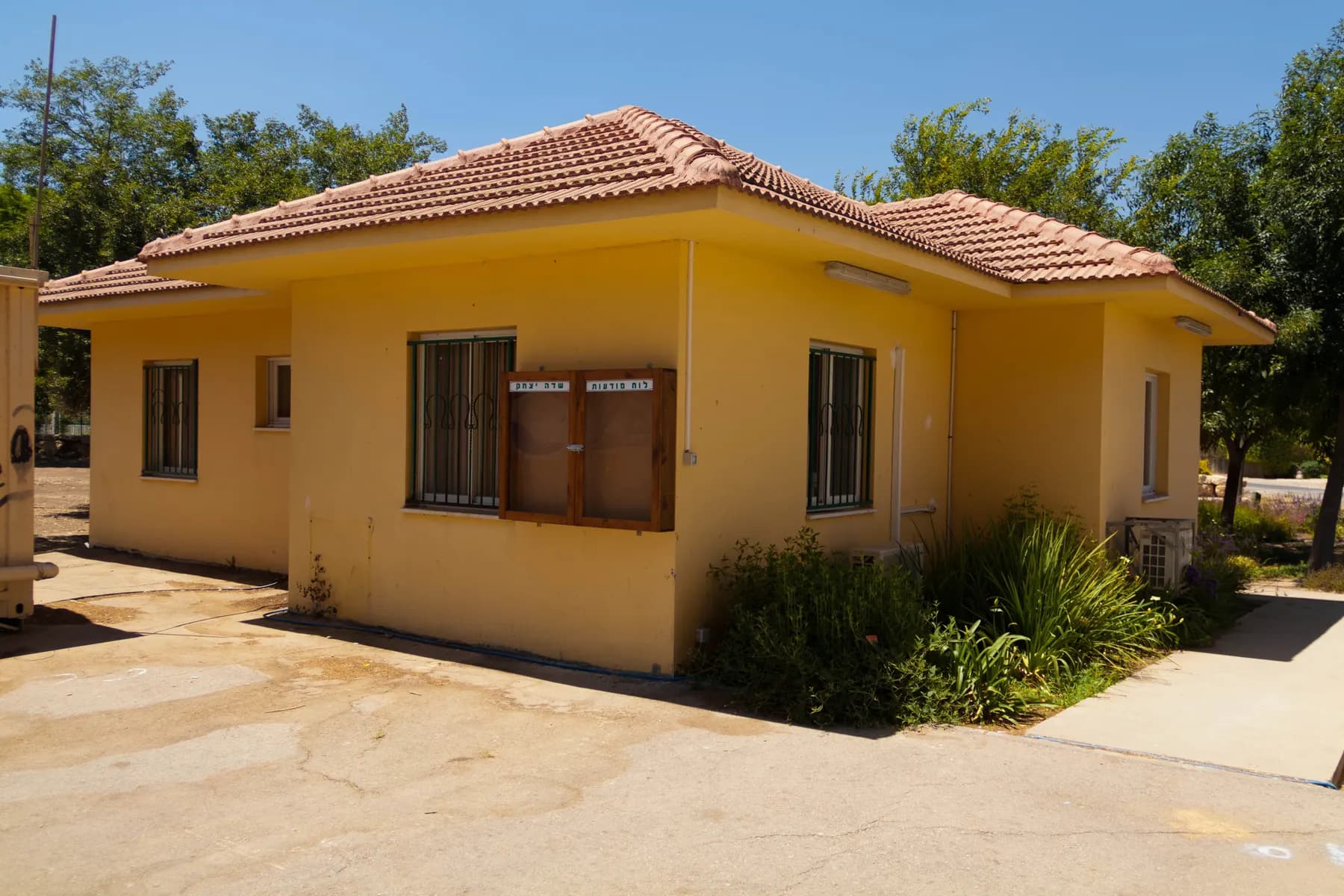The yellow single-storey office building of Sde Yitzhak, with a tiled roof, barred windows and shrubs along its wall.