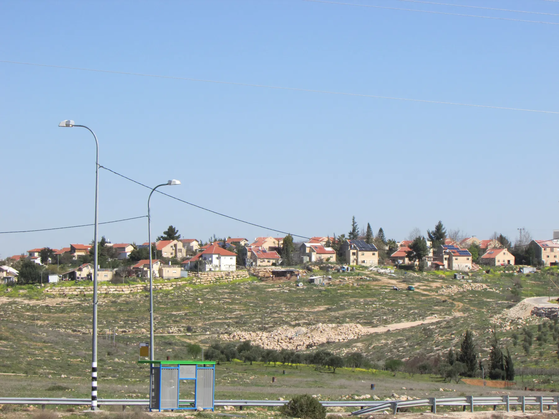 Red-roofed houses, some with solar panels, along a hilltop in Susya, above a grassy slope with a bus shelter and street lamps by the road.