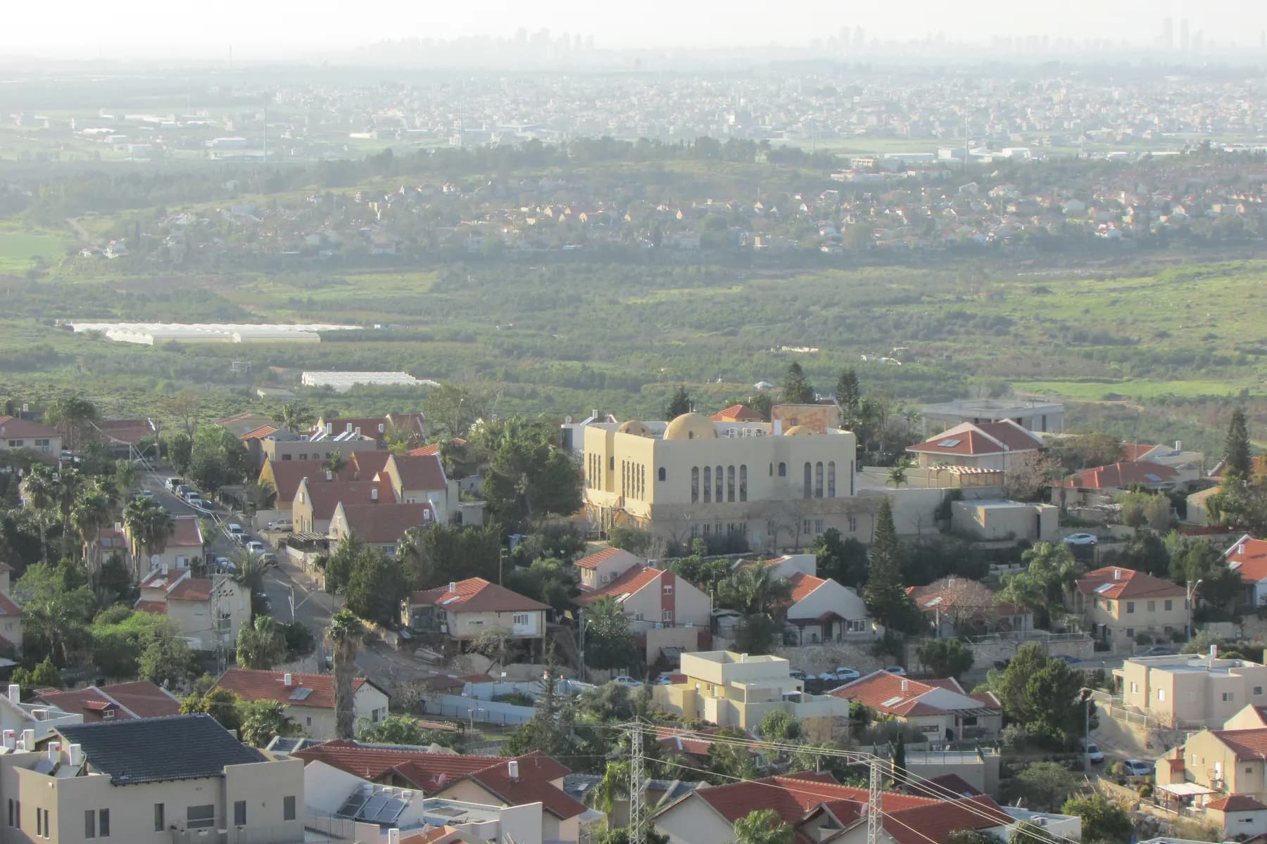 Houses with red and grey roofs and a large stone-clad building in Tzofim, seen from above, with fields and towns of the coastal plain in the haze beyond.