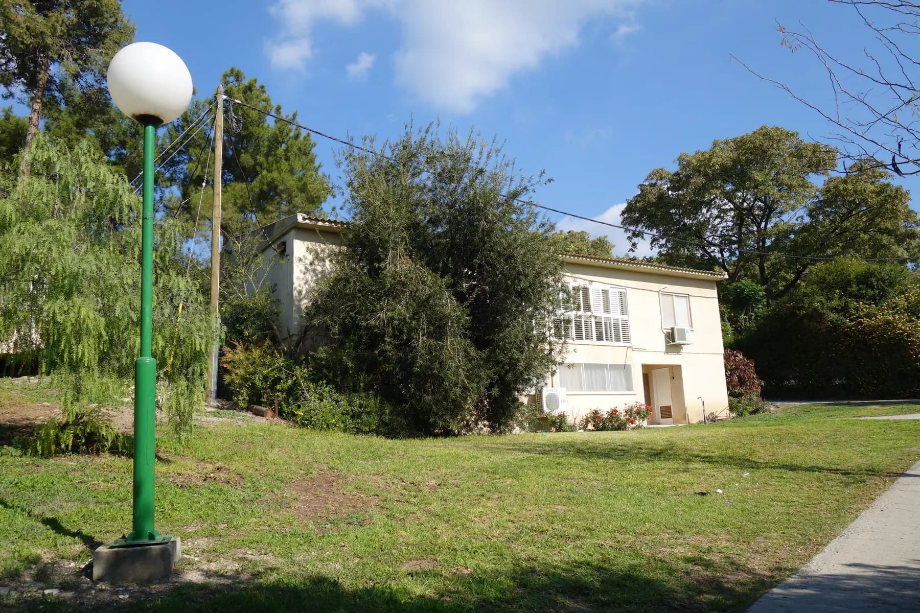 A white two-storey house behind trees on a lawn in Tzora, with a green lamp post in the foreground under a blue sky.