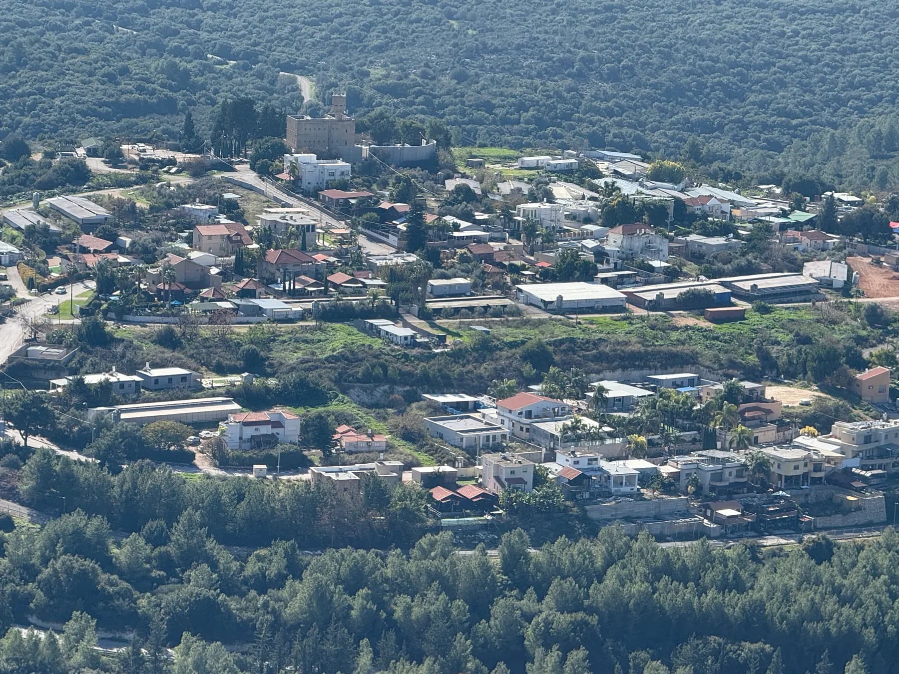 A view from a ridge over Ya'ara, with houses and farm sheds among trees, an old fort at the top of the village and woodland all around.