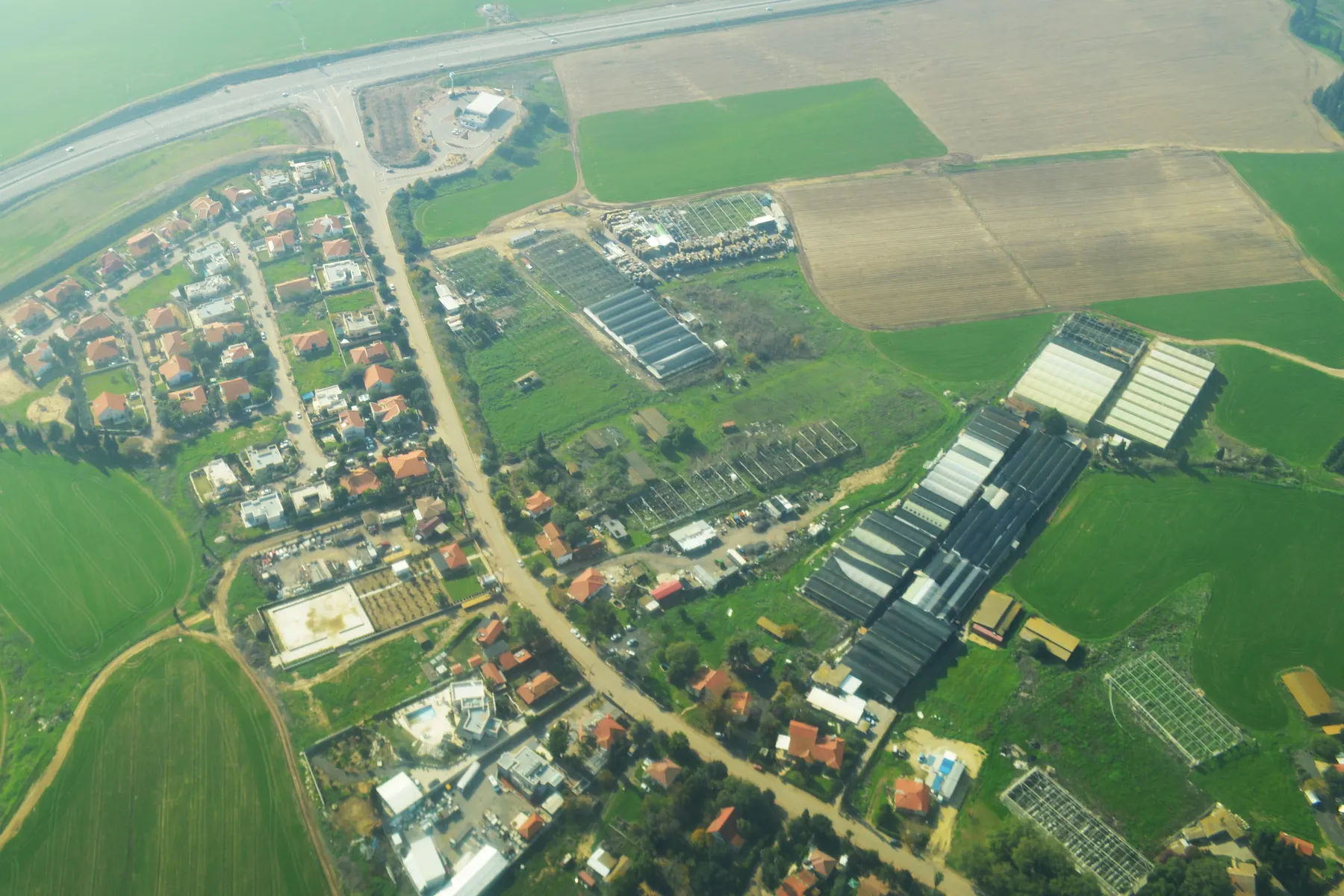 An aerial view of Yad Natan, with red-roofed houses along its roads, long greenhouses and farm sheds, and green and brown fields around them.