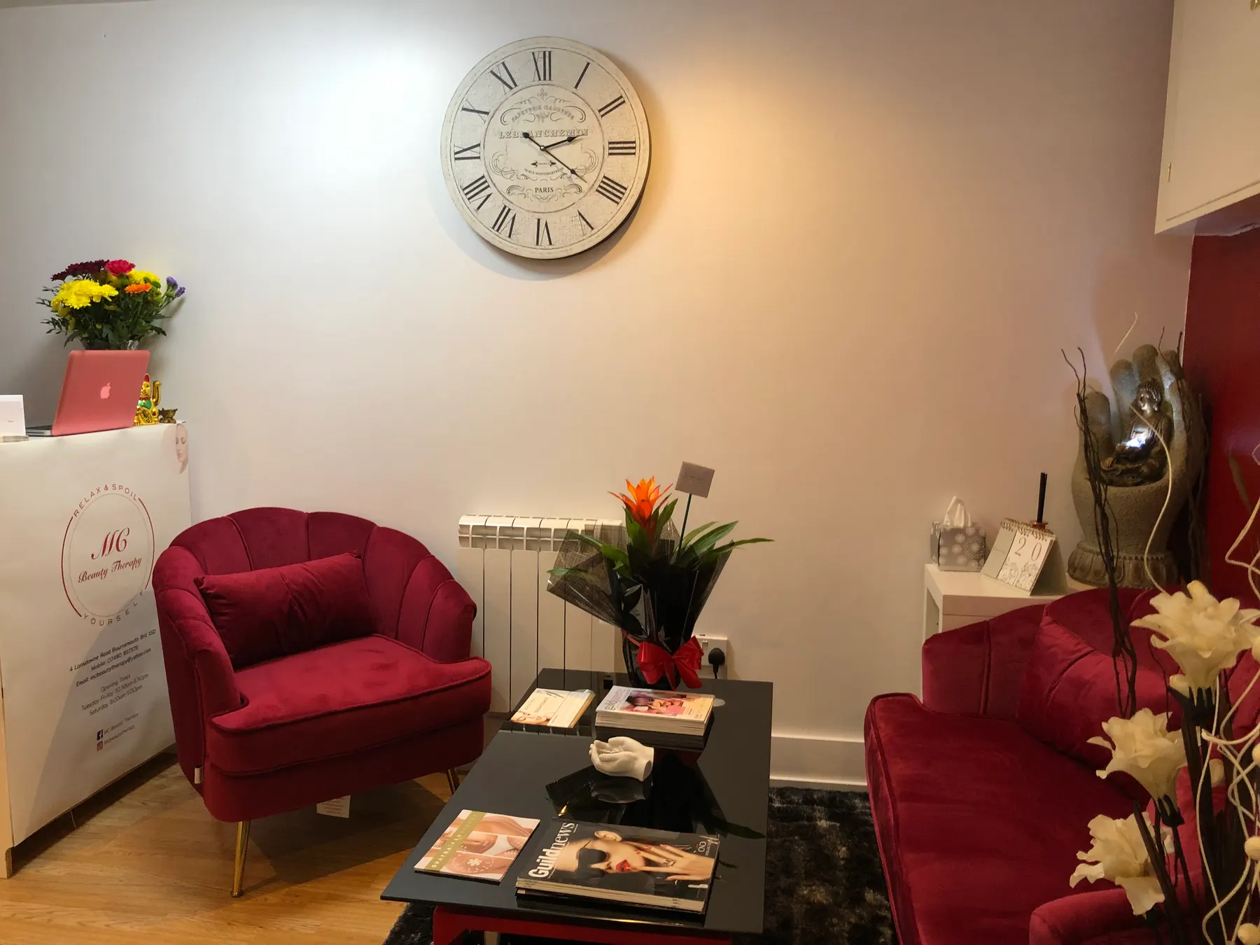 The waiting corner of a beauty salon, with red velvet armchairs, a low table, flowers and a wall clock