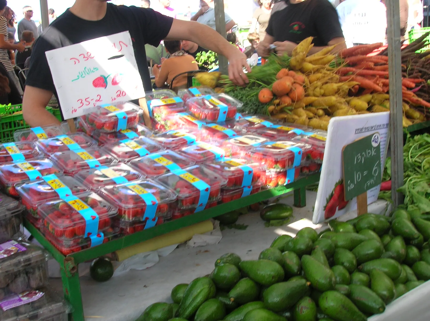 A farmers' market stall of strawberries, carrots and avocados under handwritten Hebrew price signs