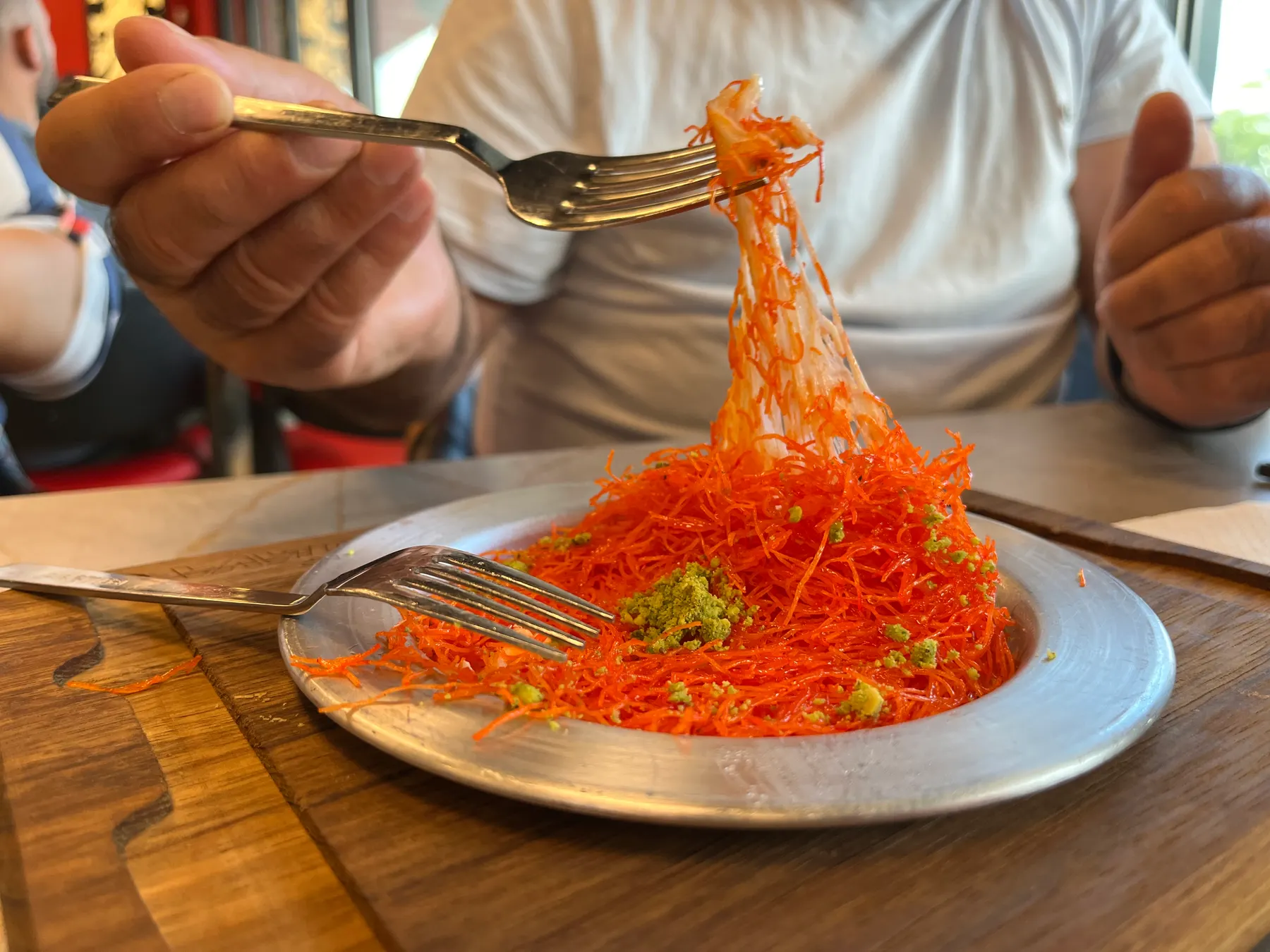 A plate of kunafa, its orange shredded pastry lifted on a fork, with pistachio on top.