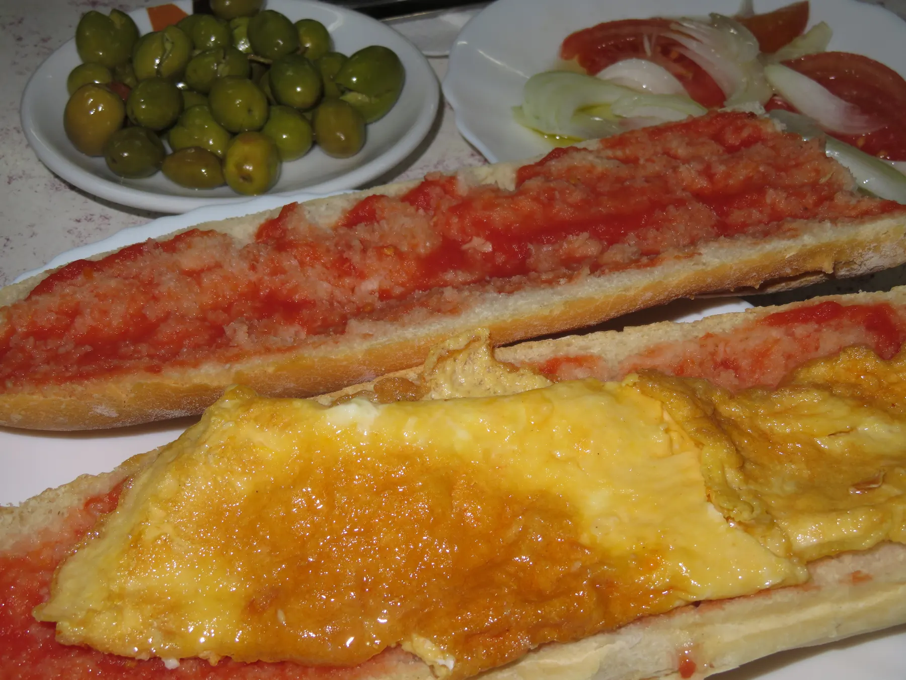 An omelette laid in a split baguette, with olives, tomato and onion on the side