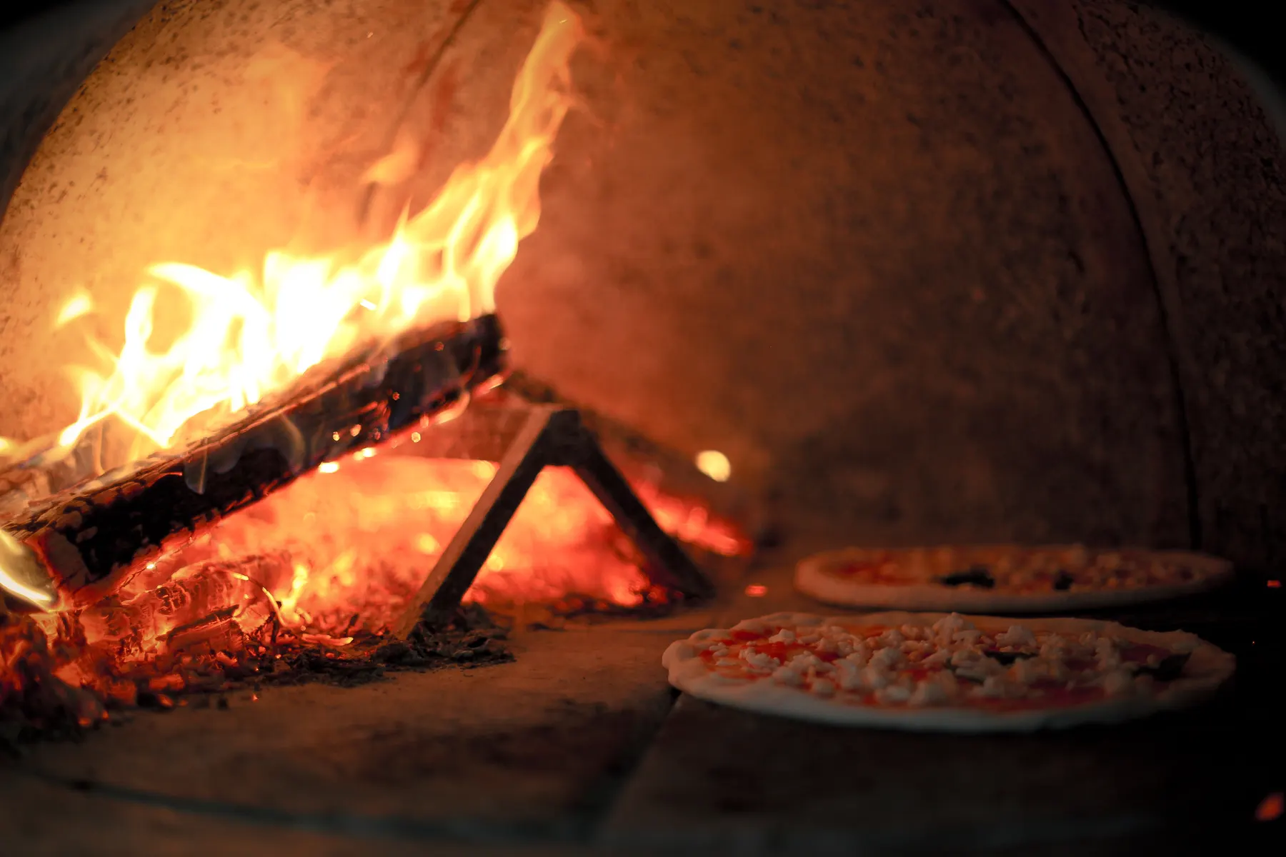 Two pizzas baking on the floor of a wood-fired oven, beside the burning logs