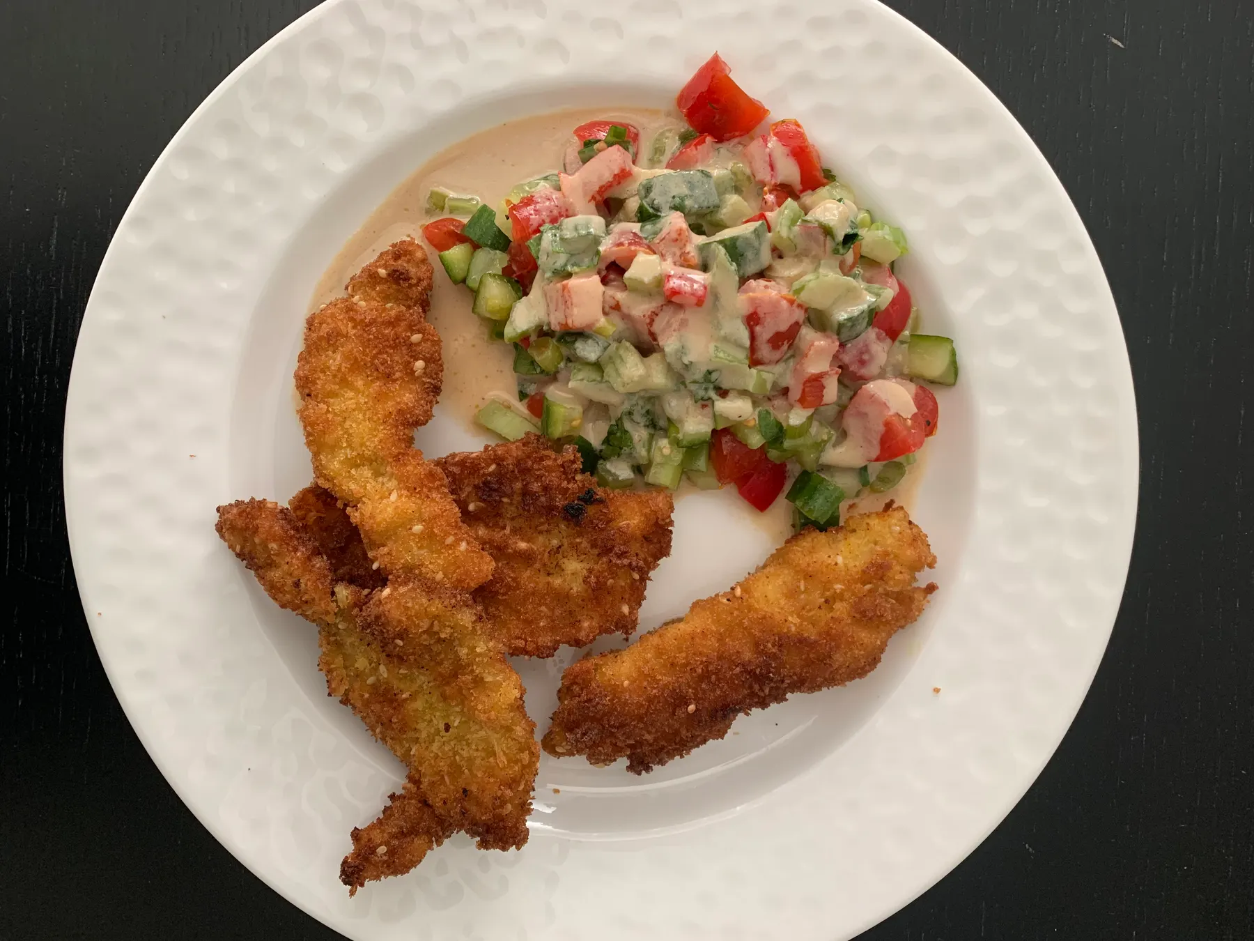 Strips of golden breaded chicken schnitzel on a white plate, with chopped Israeli salad in tahini