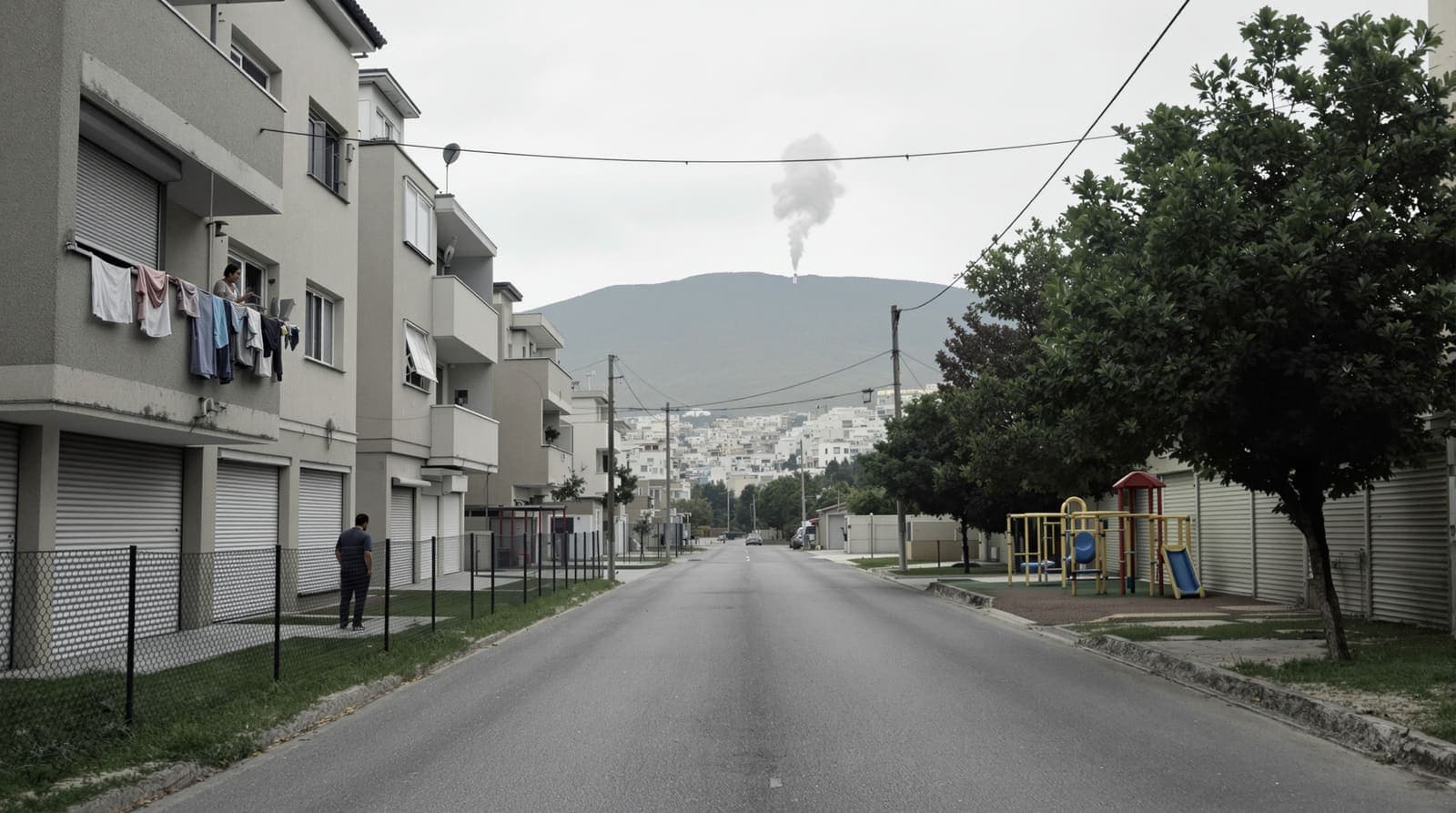 Illustration of a nearly empty Kiryat Shmona street with shuttered apartment blocks, a resident seen from behind on a laundry-hung balcony, an empty playground beside the road, and a smoke plume on a distant ridge.
