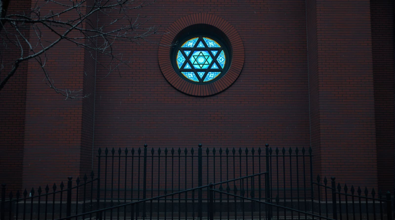Illustration of a circular stained-glass Star of David window set high in a red-brick synagogue wall above a wet Manchester street.